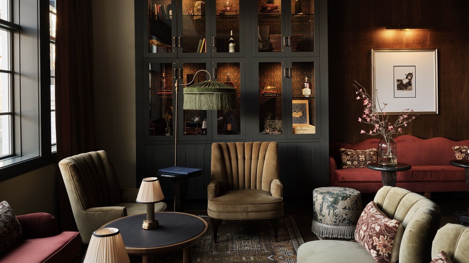 Cozy living room with vintage furniture, including a red sofa, armchairs, and patterned cushions. A dark cabinet holds various decorative items.