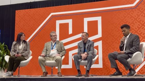 Four people sit on stage in a panel discussion. The backdrop is orange with a geometric pattern and a large letter "H".