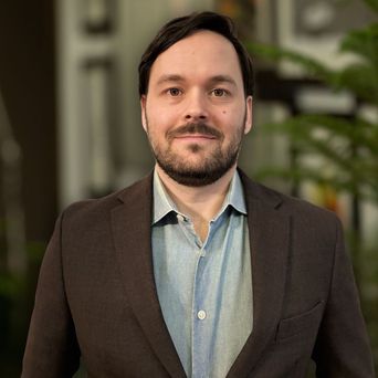 Man with short hair and a beard wearing a brown blazer over a blue shirt, standing indoors with plants and a lamp in the background.