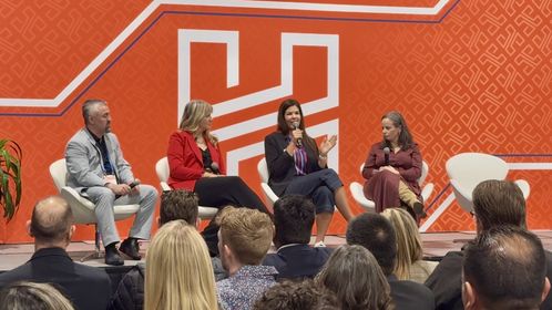 Four people sit on stage in a panel discussion against an orange backdrop, with an audience in the foreground.