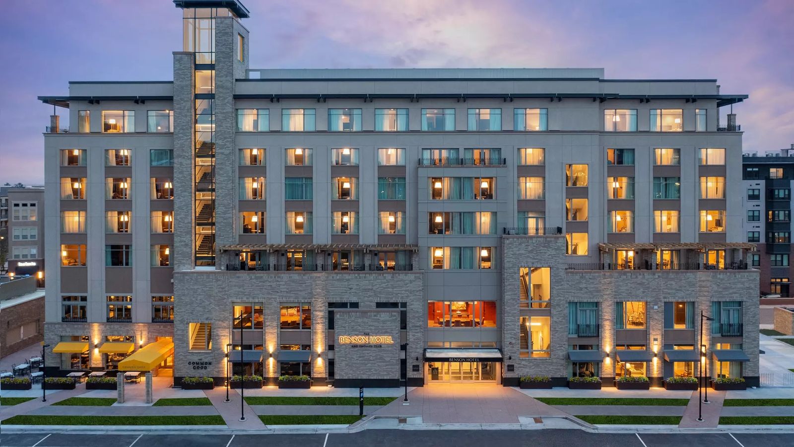 Modern multi-story hotel at dusk, with warm lit windows, stone and glass facade, and an inviting entrance with surrounding urban street.