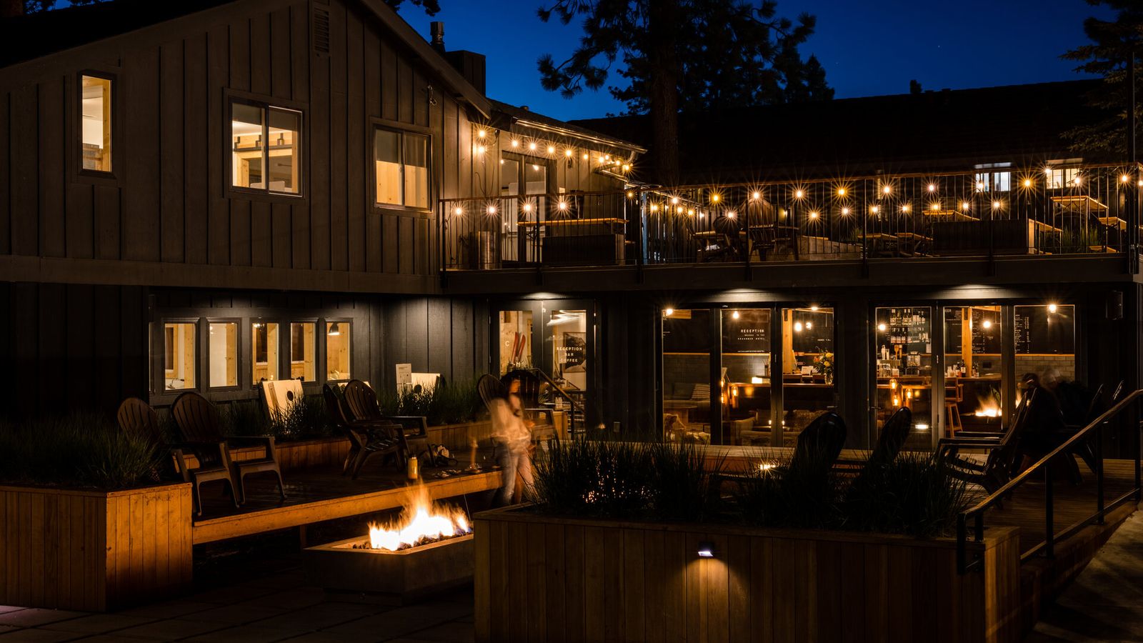 Cozy outdoor patio at night with a fire pit, string lights, and wooden seating, adjacent to a modern building with large windows.