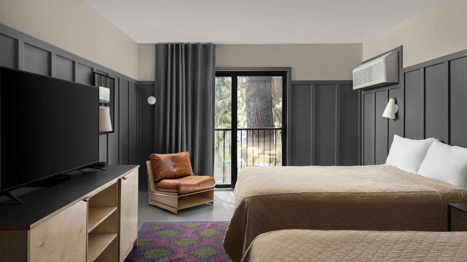 Modern hotel room with two beds, a TV, a brown leather chair, and a balcony with a view of trees. The decor features dark paneling and a colorful rug.