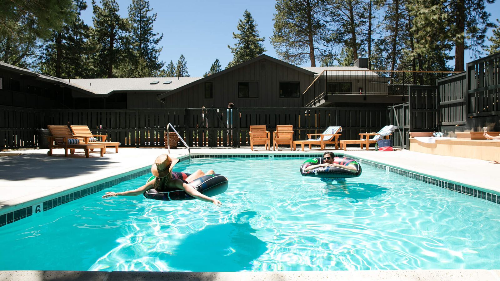 Two people relax on inflatable rings in a sunlit outdoor pool, surrounded by trees and wooden loungers, under a clear blue sky.
