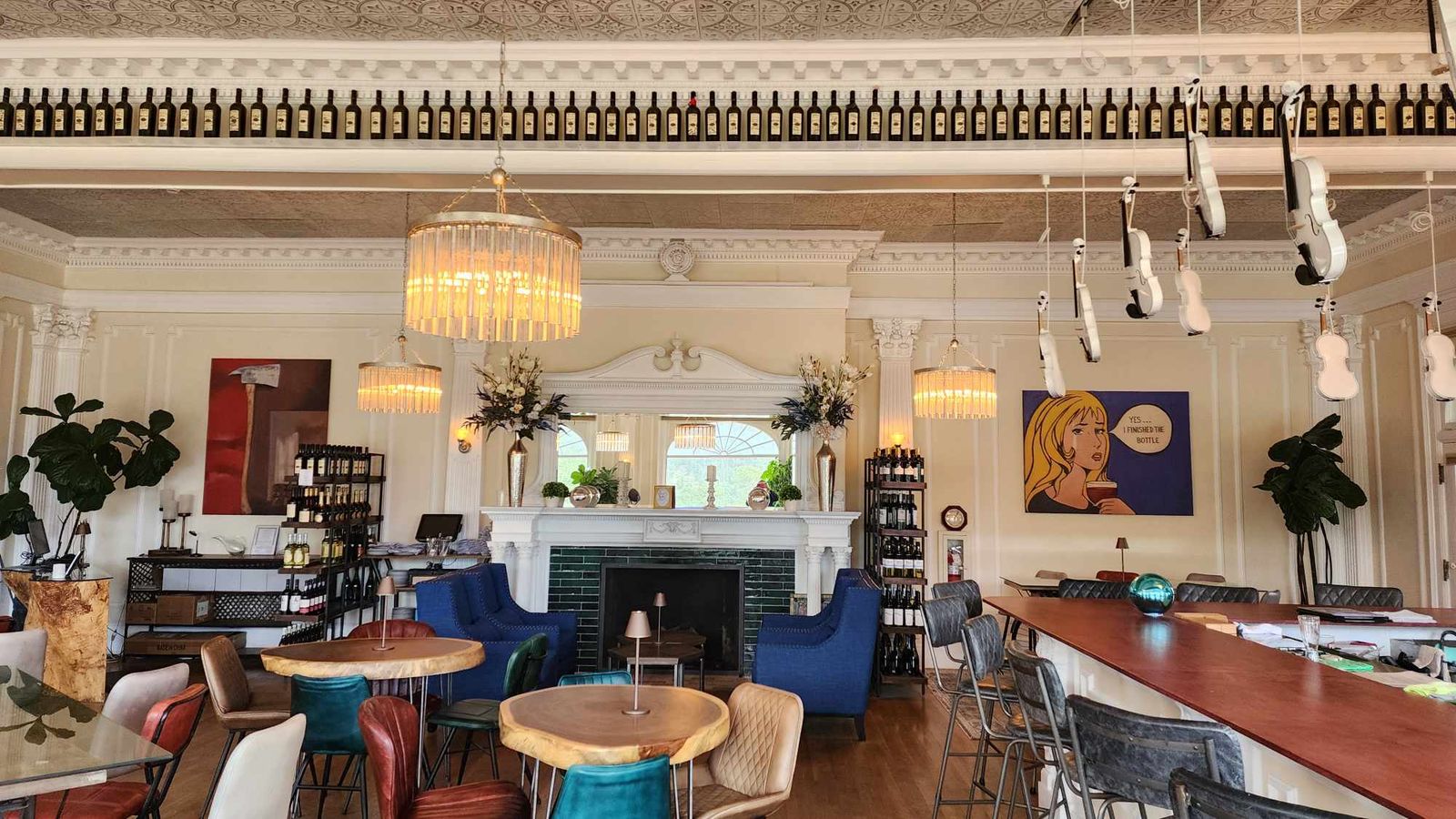 Stylish cafe interior with eclectic seating, chandeliers, wall art, and wine bottles displayed above. Cozy and inviting atmosphere.