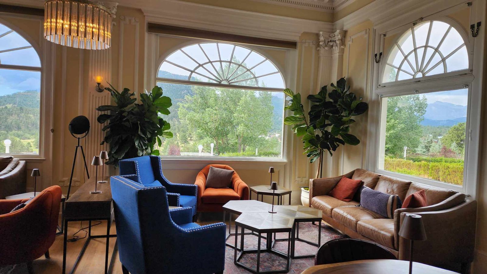 Elegant lounge with large windows, blue and brown chairs, leather sofa, and lush plants, overlooking a scenic view of trees and mountains.