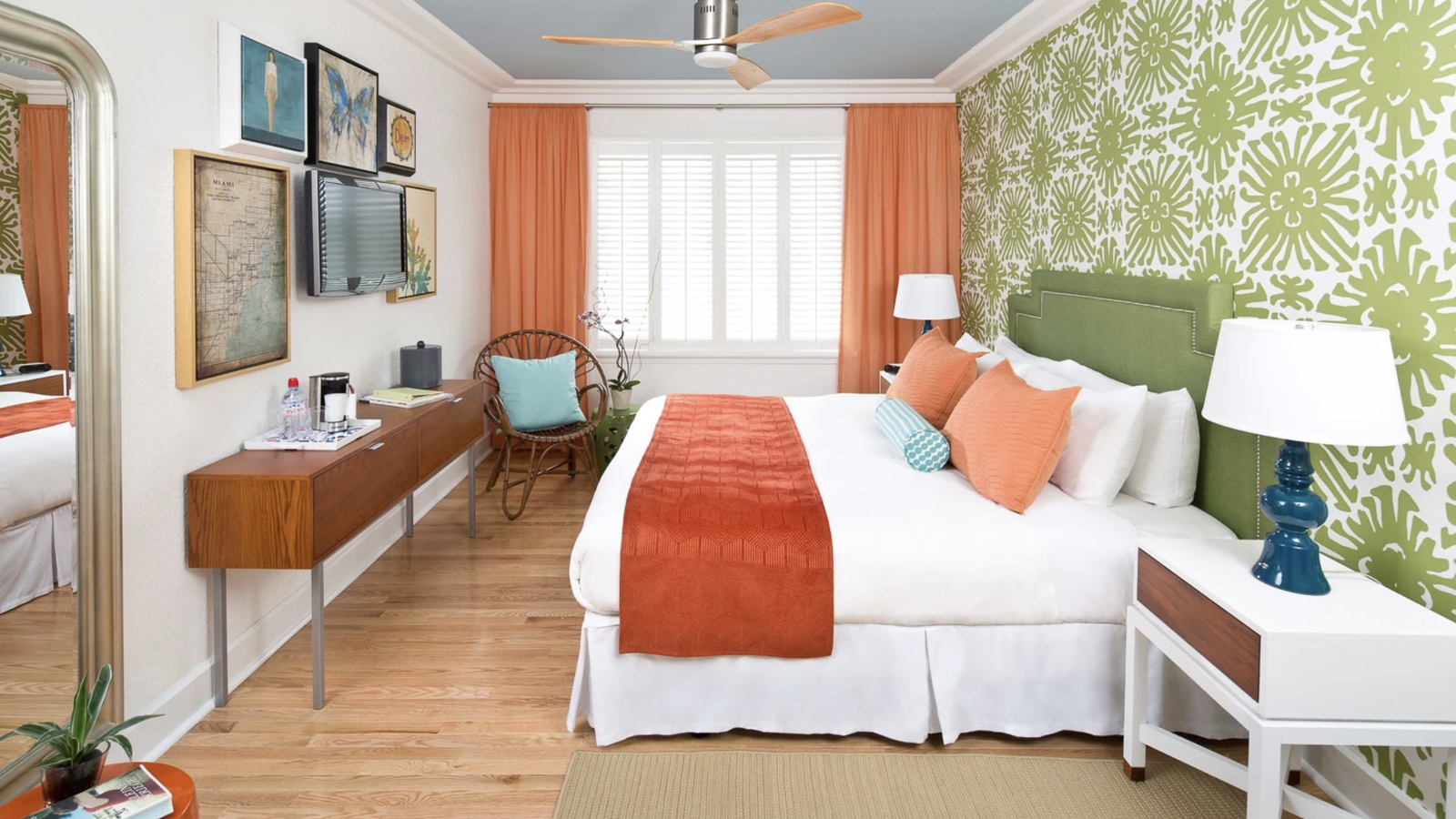 Bright bedroom with green and white patterned wall, orange accents, a bed with colorful pillows, wooden floor, and modern furniture.