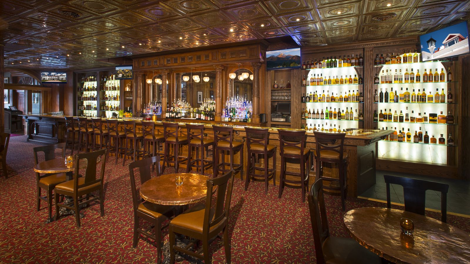 Elegant bar with wooden decor, high chairs, round tables, and illuminated shelves filled with bottles, set on a red patterned carpet.