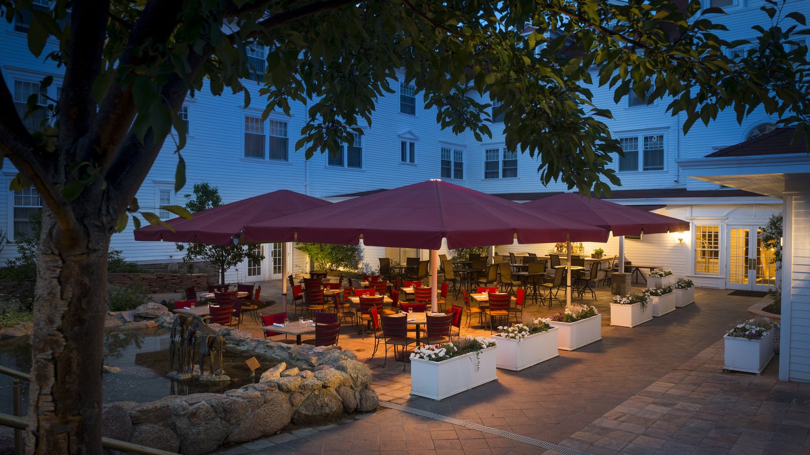 Outdoor dining area with red umbrellas and wooden tables, surrounded by white planters and a stone path, under a twilight sky.