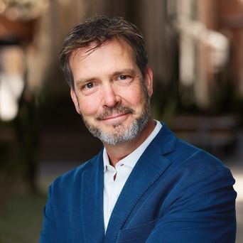 Man with short hair and beard, wearing a blue suit jacket and white shirt, smiling with arms crossed, outdoor blurred background.