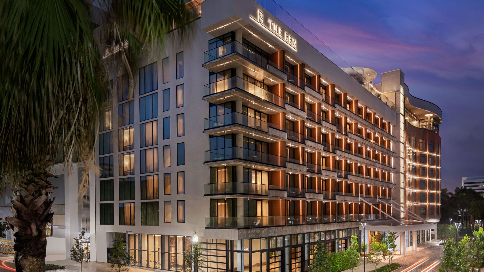 Modern hotel with illuminated windows and balconies at dusk, surrounded by greenery and lit street lamps.