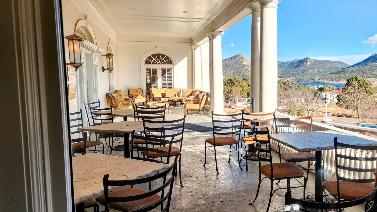 Outdoor patio with empty tables and chairs, white columns, and a view of mountains under a clear blue sky.