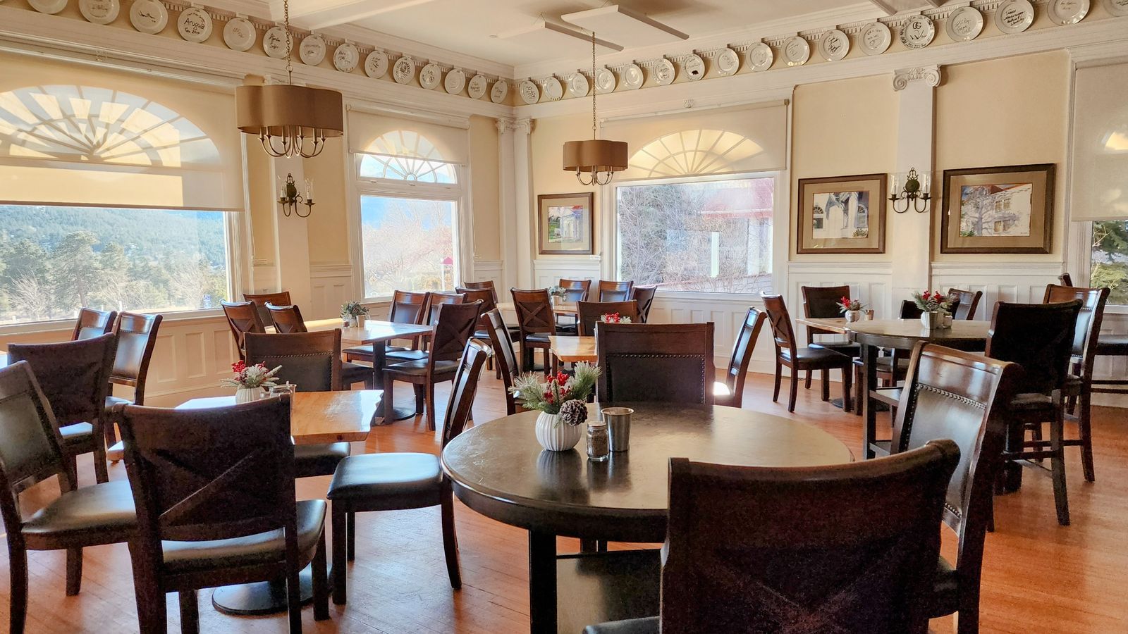 A bright, elegant dining room with wooden tables and chairs, large windows, chandeliers, and framed artwork on the walls.