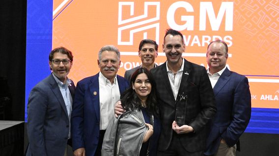 A group of six people, five men and one woman, stand together smiling in front of a backdrop with "GM Awards" text. One man holds an award.
