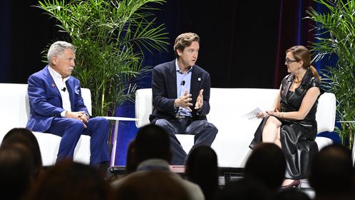 Three people seated on a stage engage in a panel discussion, with a woman holding notes. Two potted plants are in the background.
