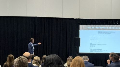 A speaker in a suit presents to an audience in a conference room, gesturing towards a screen displaying a slide titled "Use AT Tools for Work and Personal."