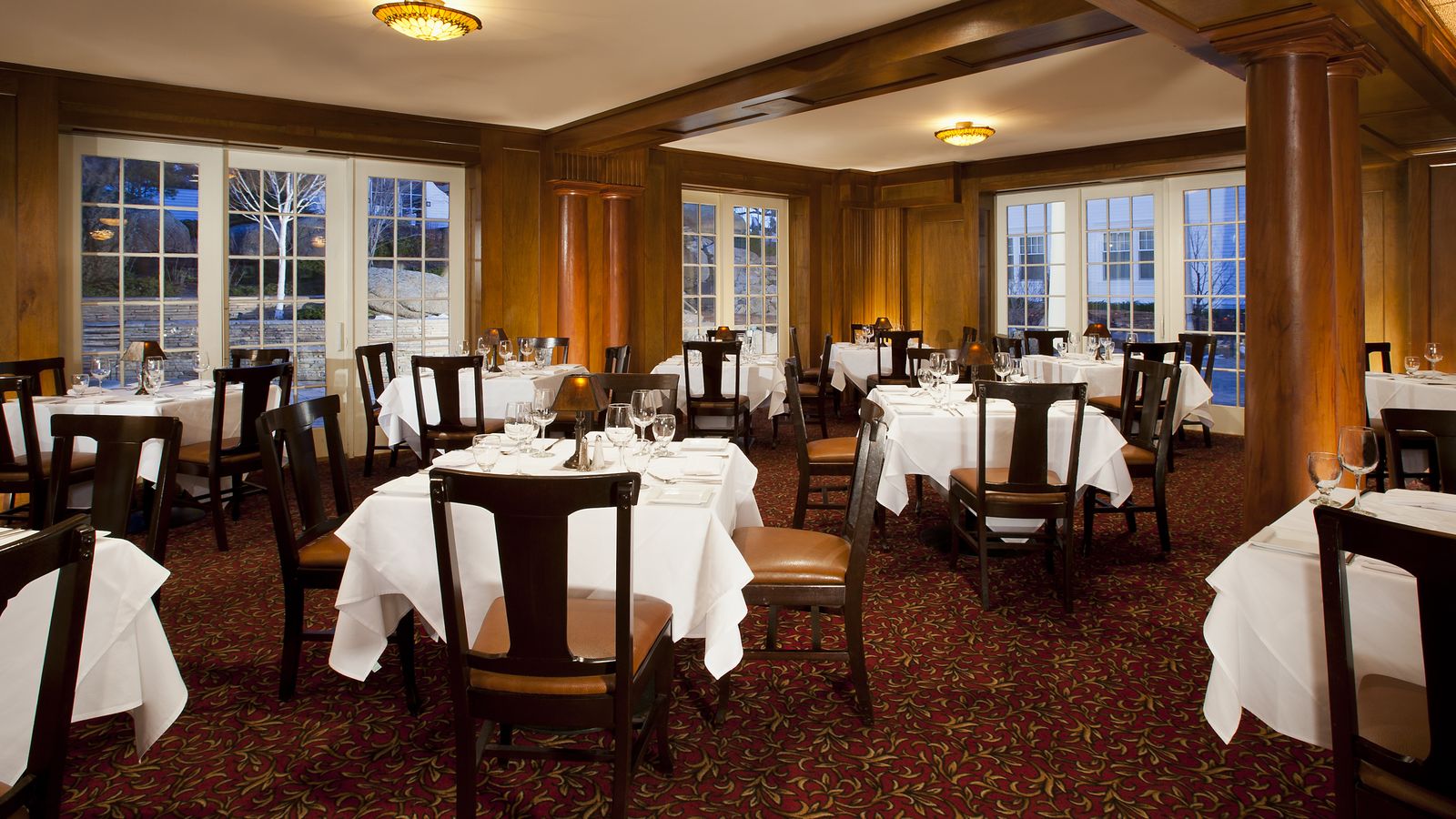 Elegant restaurant interior with wooden paneling, red patterned carpet, and tables set with white tablecloths and dining ware. Large windows in view.