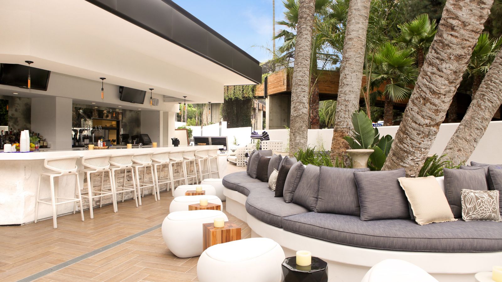 Outdoor lounge with a sleek bar, white stools, cushioned seating, palm trees, and modern decor under a clear blue sky.