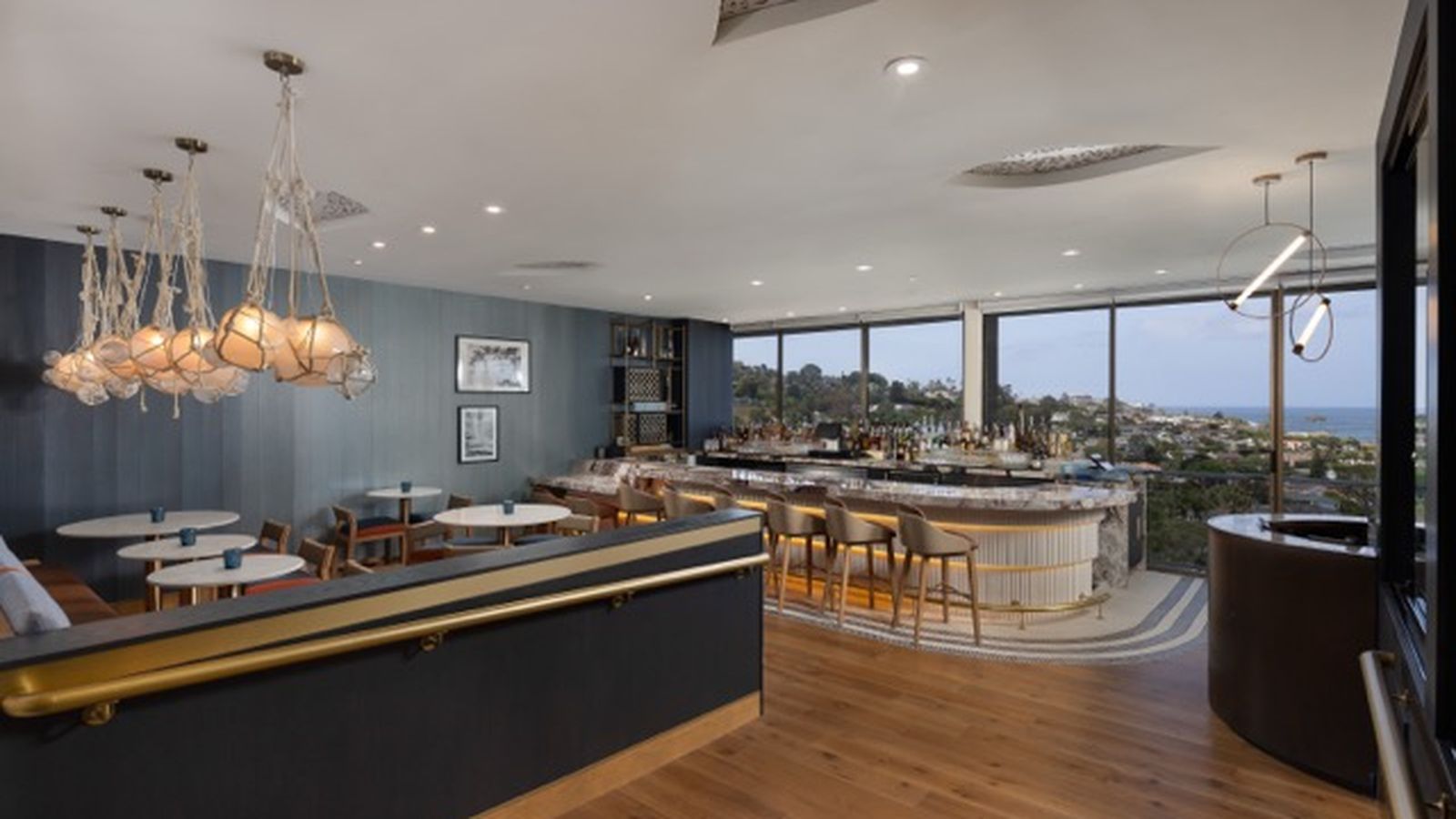 Modern bar interior with elegant lighting, round tables, and a curved bar. Large windows offer a scenic view of a hillside and coastline.