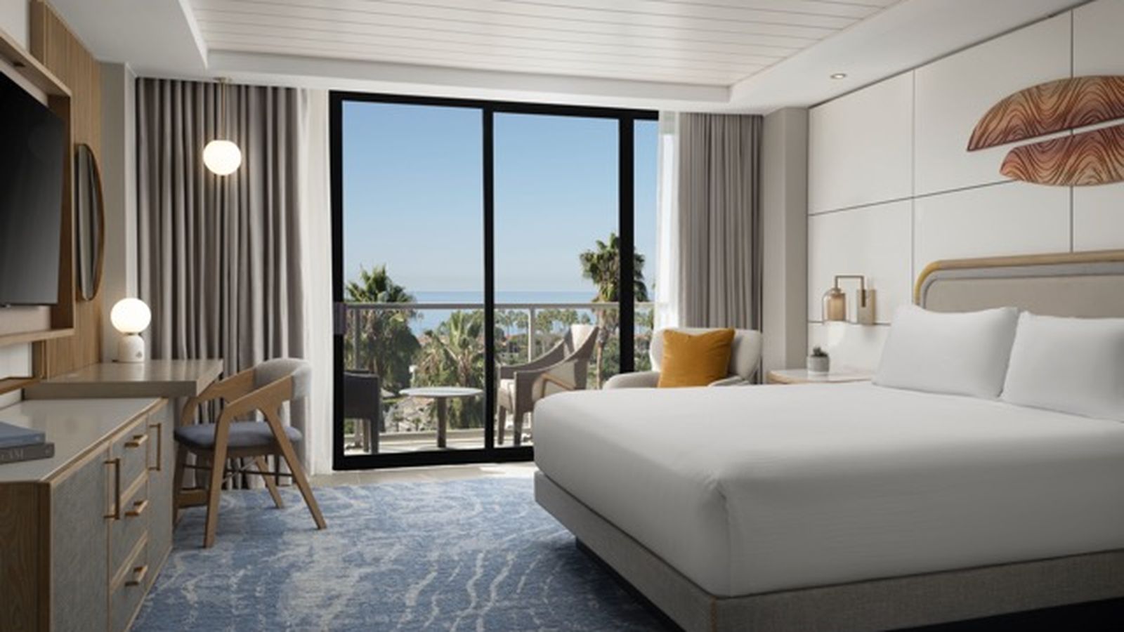 Modern hotel room with a large bed, desk, and TV. Glass doors open to a balcony with outdoor seating, offering a view of palm trees and the ocean.