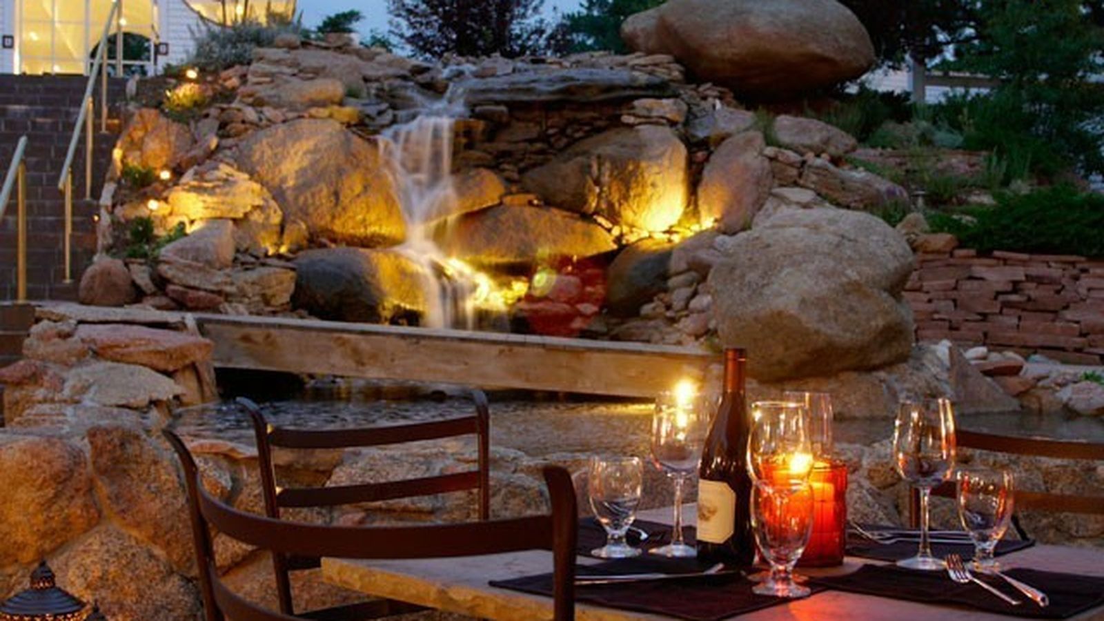 Romantic outdoor dining setup with candles and wine on a table, overlooking a lit waterfall and rocks in an evening garden setting.