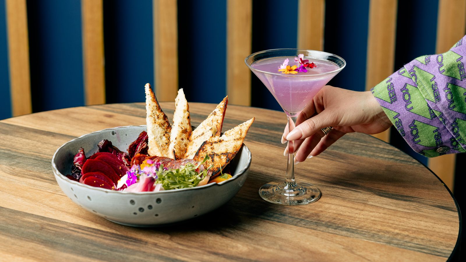 A hand reaches for a purple cocktail in a martini glass, garnished with edible flowers, next to a ceramic bowl filled with vibrant vegetables, toasted bread slices, and colorful garnishes, all set on a wooden table with a blue and wood slat background.