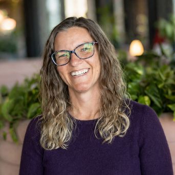 Smiling woman with wavy hair and glasses, wearing a purple sweater, standing in front of greenery with a blurred background.