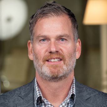 Man with grey beard, wearing a patterned suit jacket and checkered shirt, smiling with a blurred indoor background.