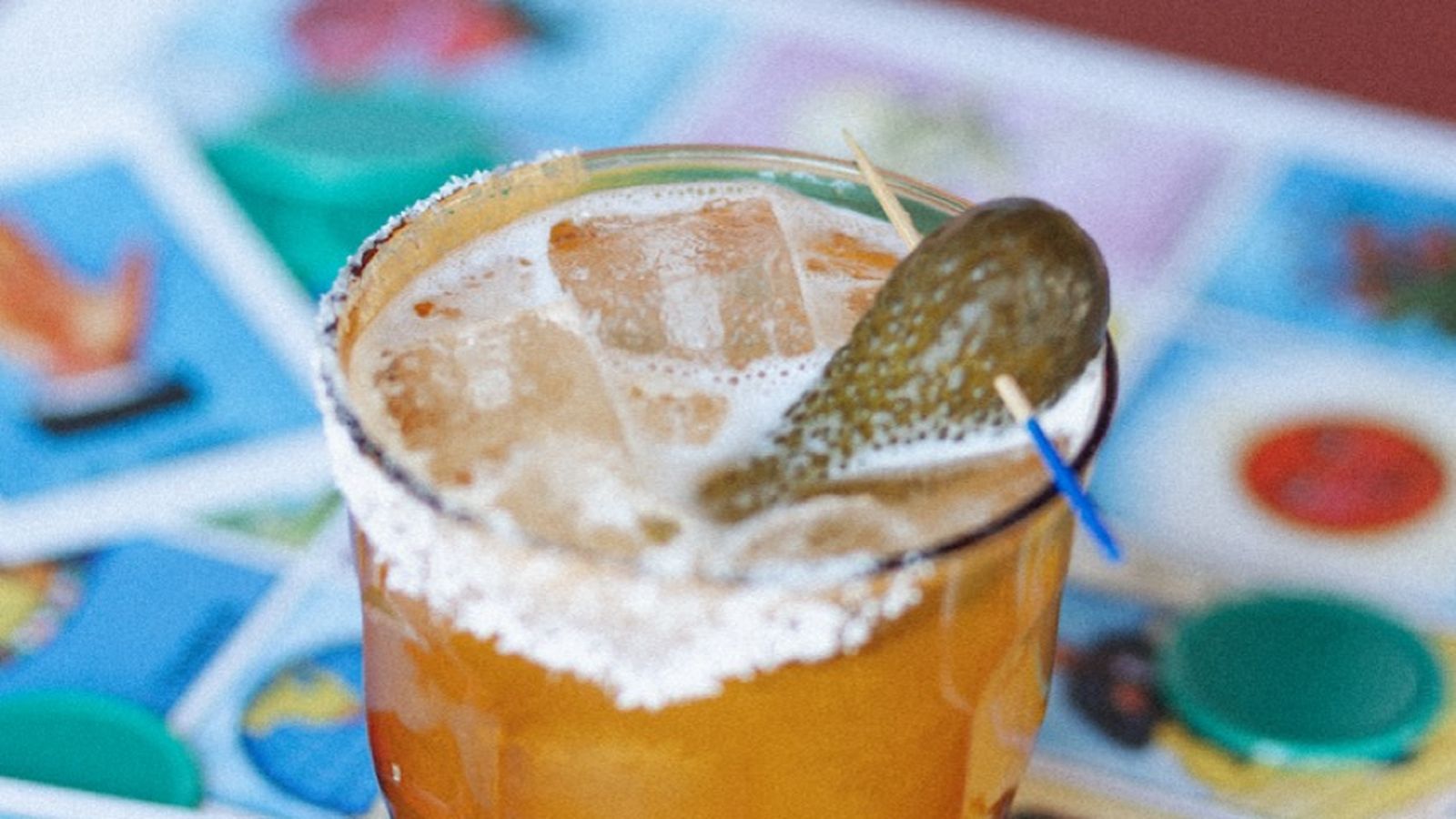 A cocktail with a salted rim, ice, and a pickle garnish on a red surface, placed over colorful cards.