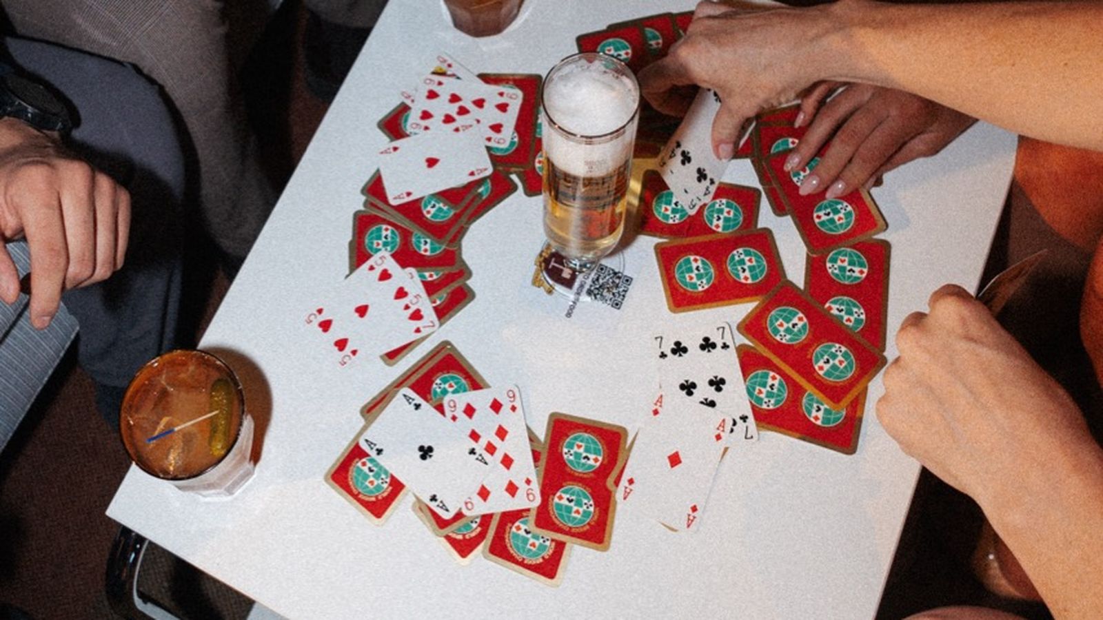 People playing cards at a table with drinks, scattered cards, and a wallet.