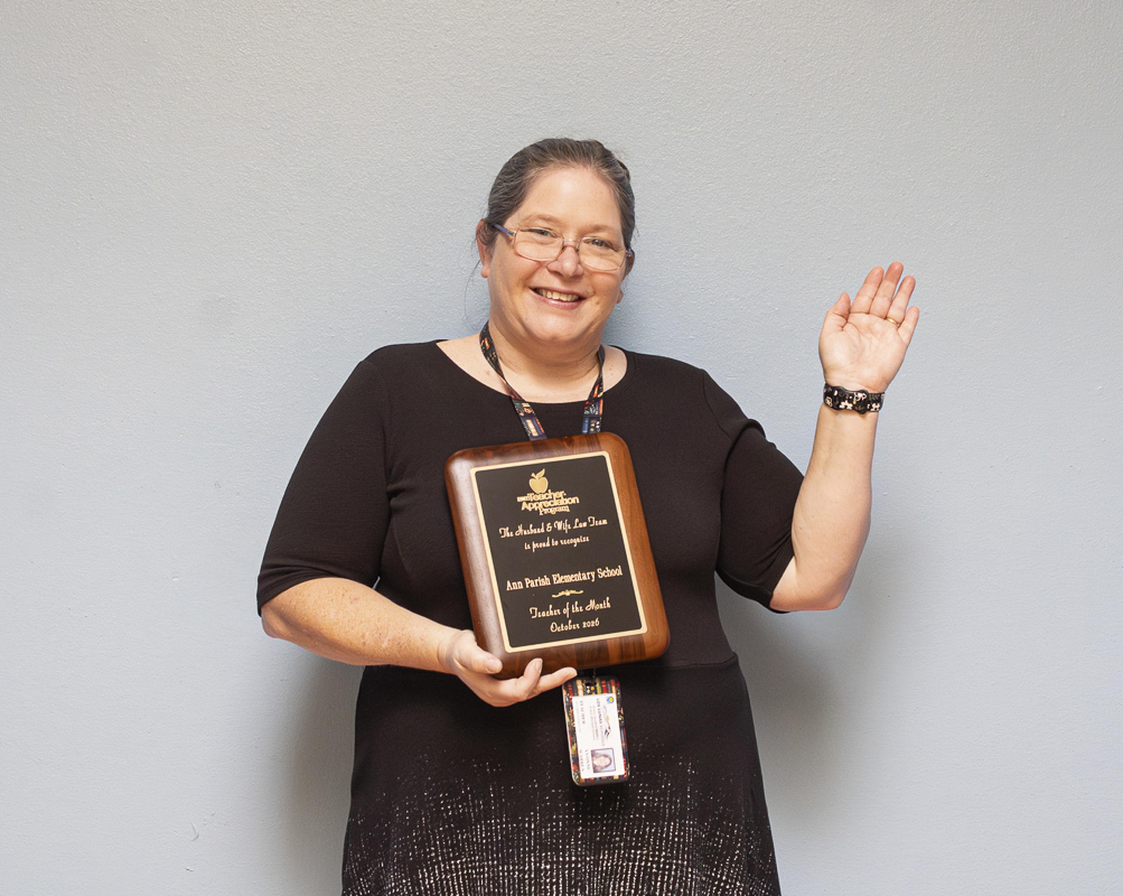 Mrs. Warren smiles and waves while proudly displaying her Teacher of the Month award at Ann Parish Elementary School.