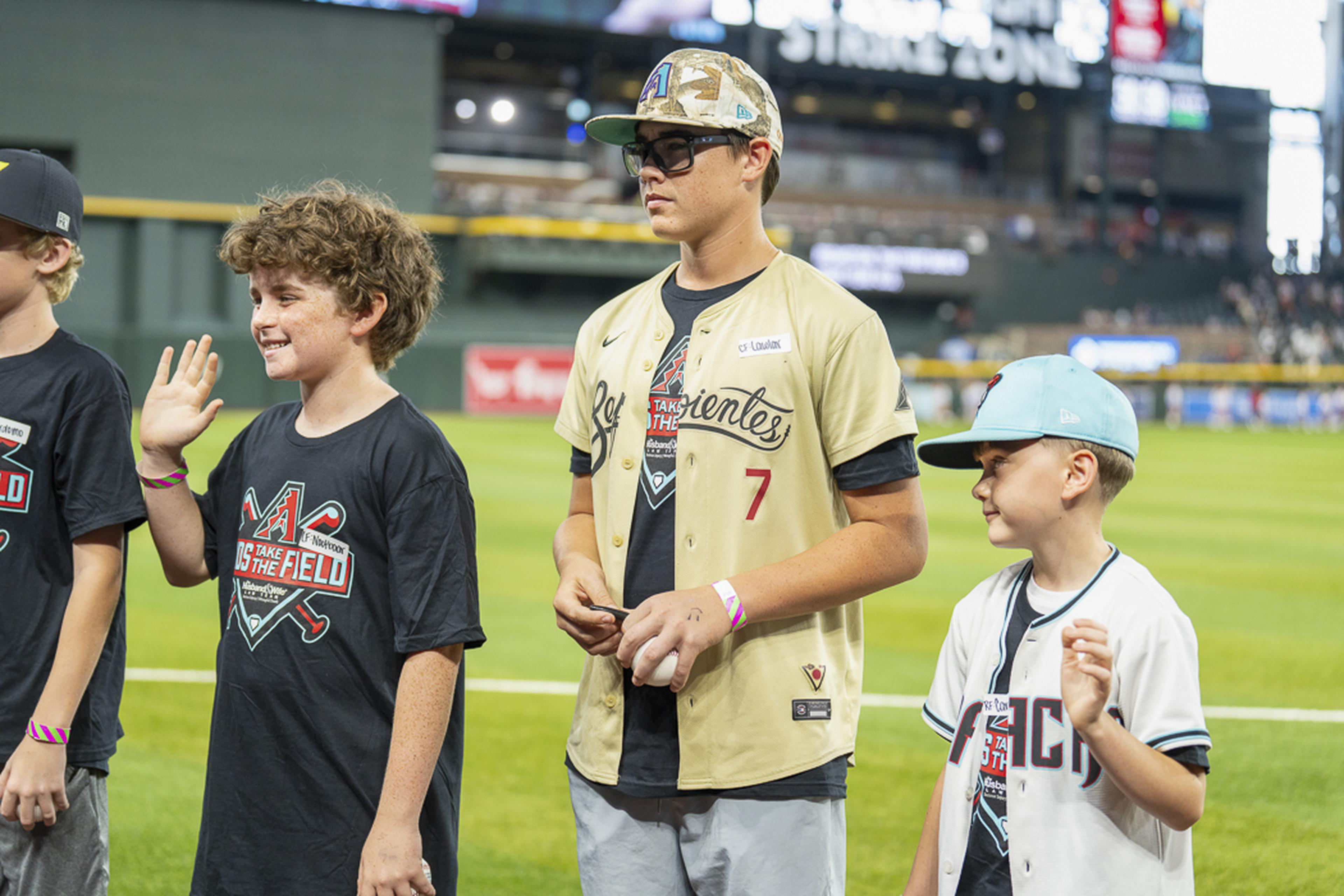 Three young Kids Take the Field participants smile and wave from the field, sharing their excitement during a memorable day at Chase Field before the game.