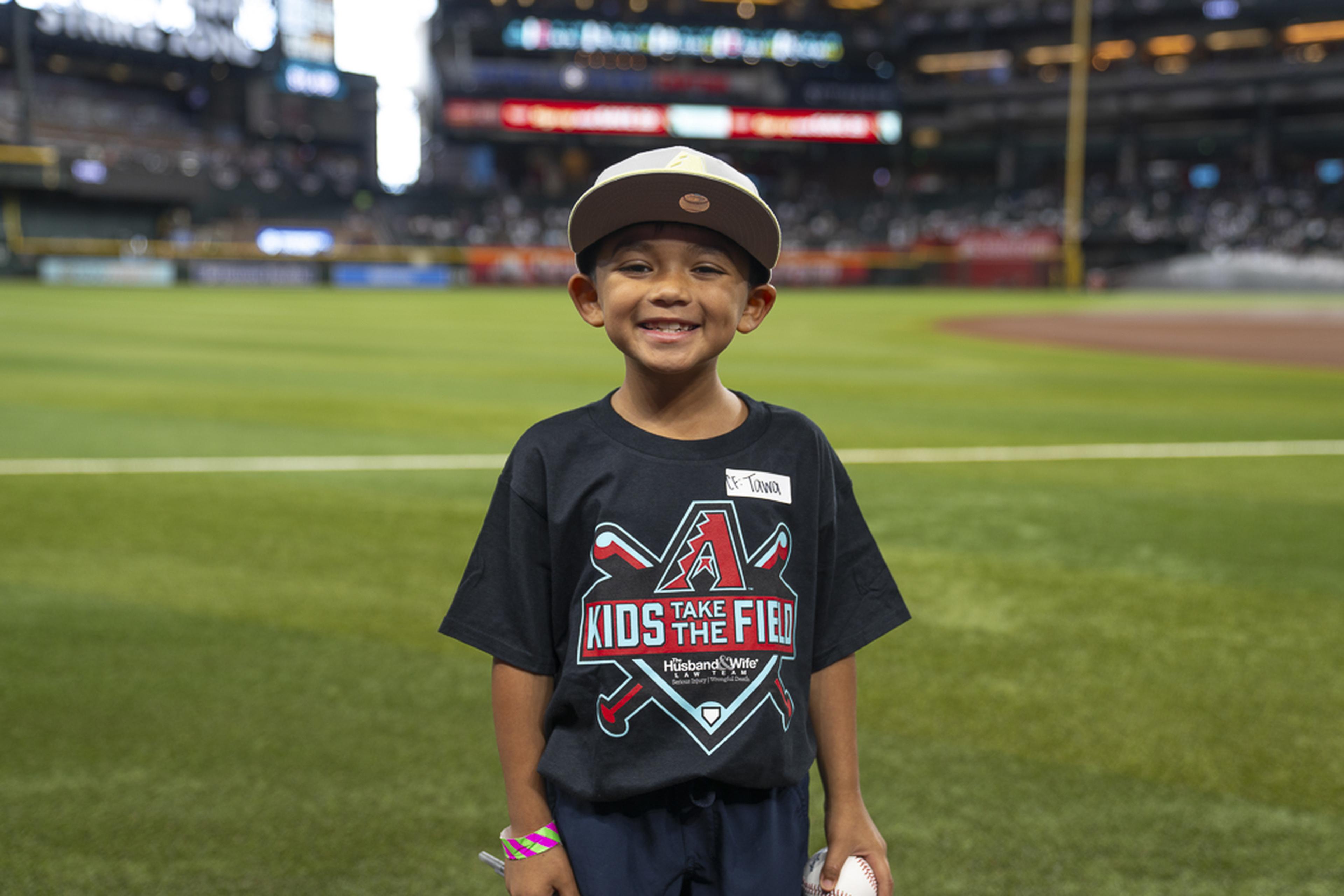 A young Kids Take the Field participant proudly holds a baseball on the field, smiling as he enjoys a memorable day at Chase Field with the excitement of game day all around him.