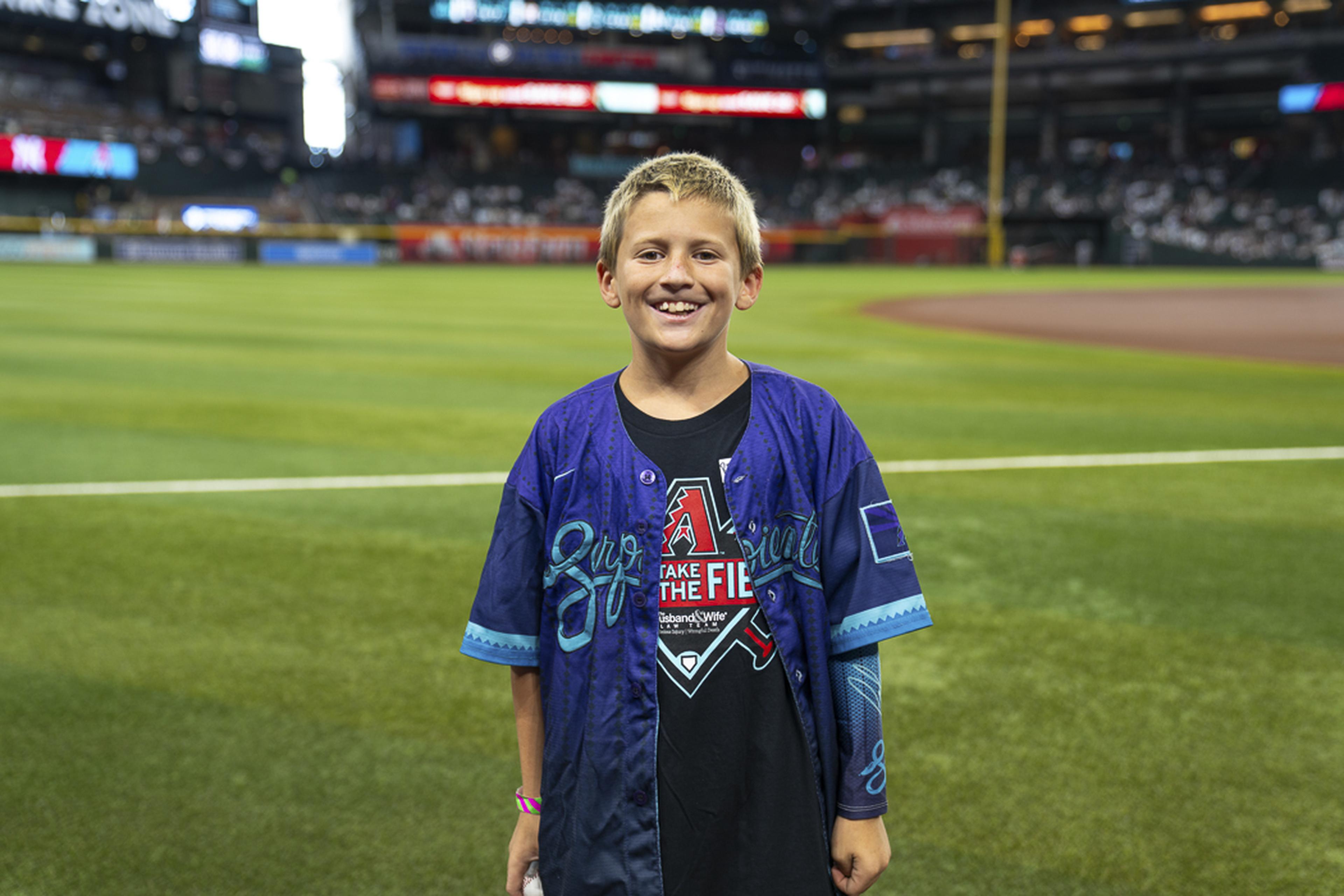 A young Kids Take the Field participant smiles on the field at Chase Field, taking in the excitement of game day with thousands of fans in the stands behind him.