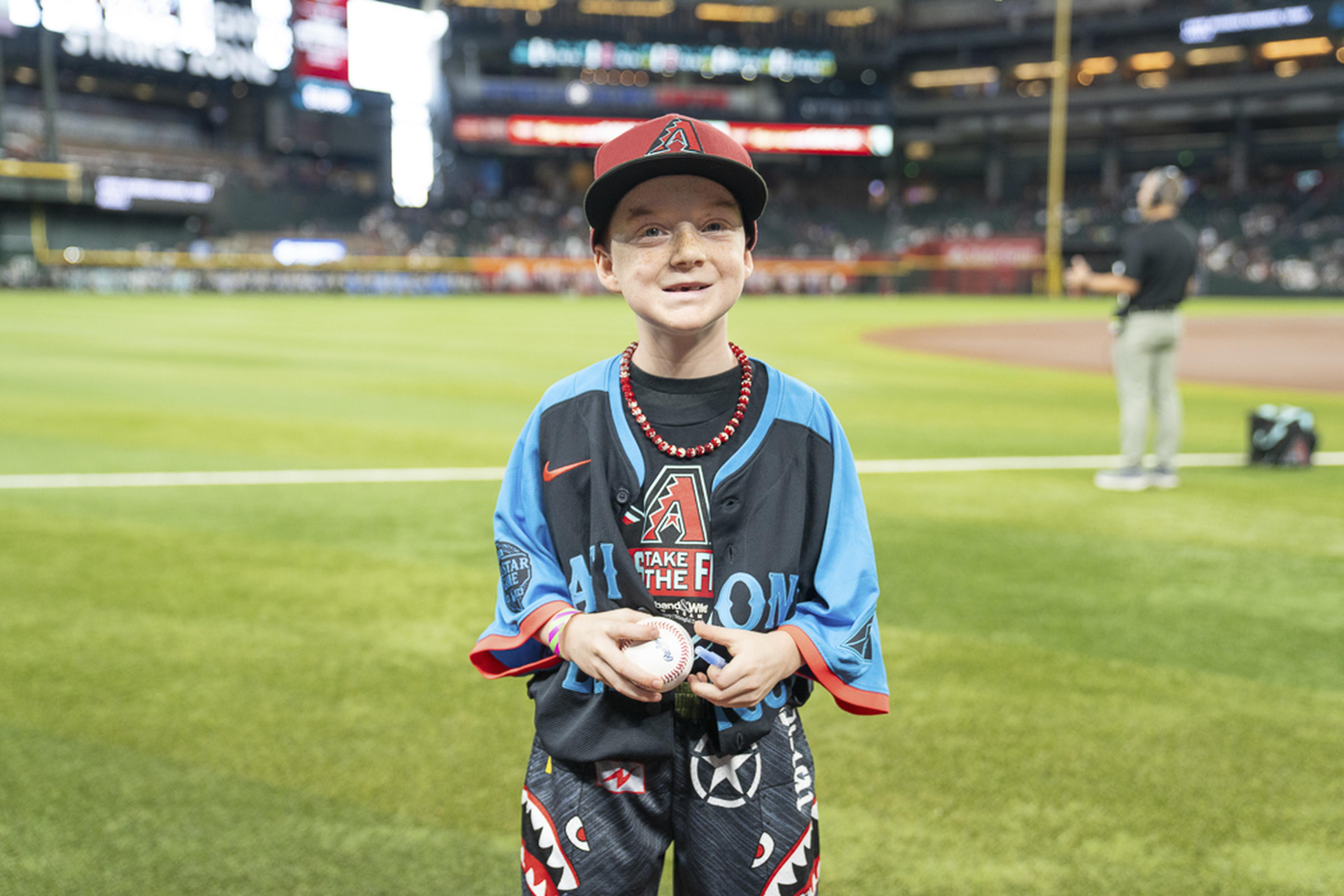 A young fan proudly holds a baseball on the field, soaking in the excitement of his Kids Take the Field experience at Chase Field with the stadium crowd behind him.