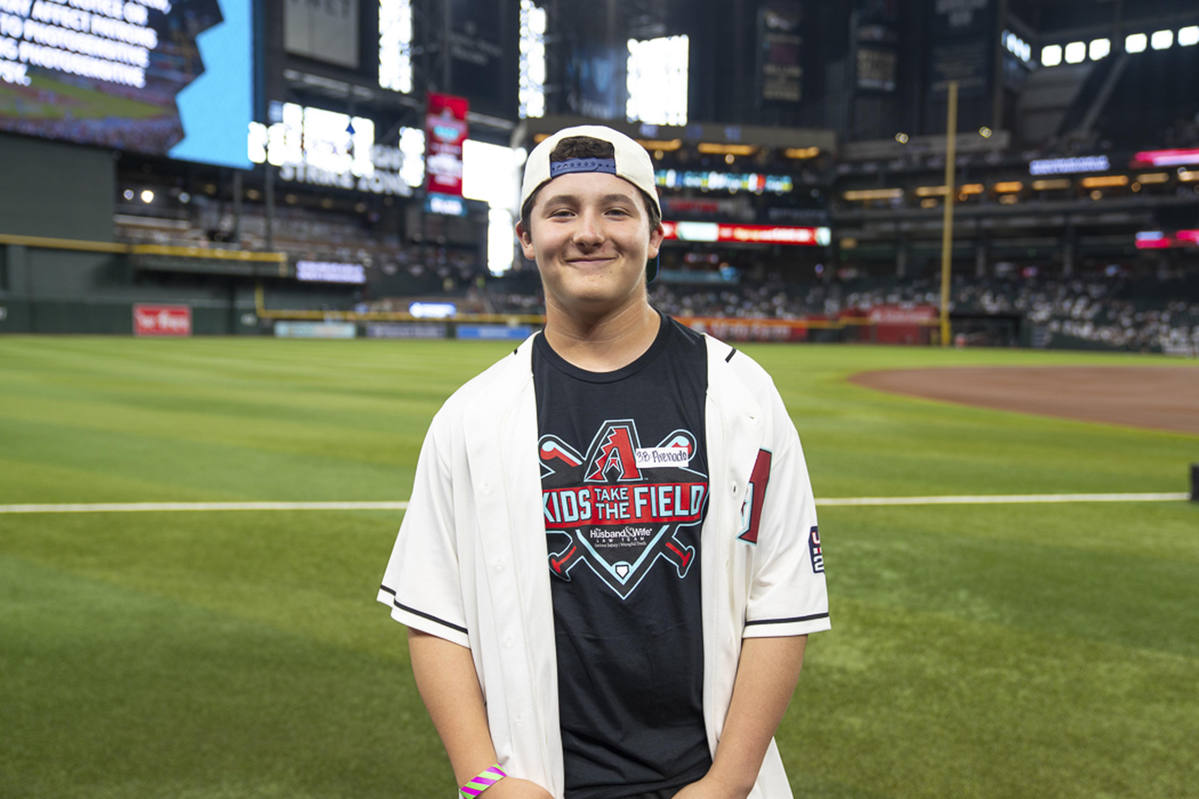 Wearing a Kids Take the Field shirt and a backward cap, a young fan smiles on the field as he enjoys a once-in-a-lifetime experience at Chase Field.