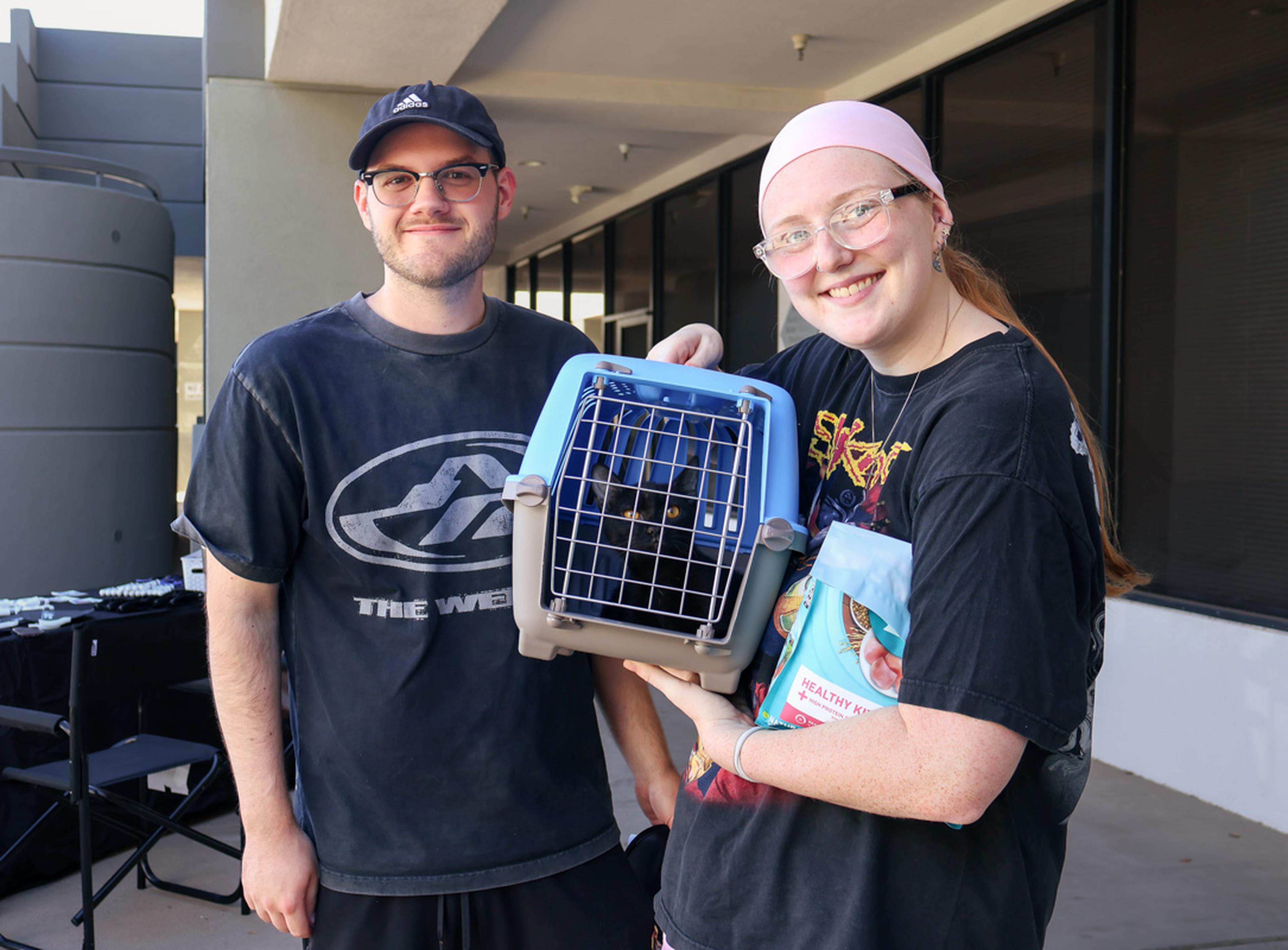 Two people smiling, holding a pet carrier with a black cat inside. One person is holding a bag of pet food.