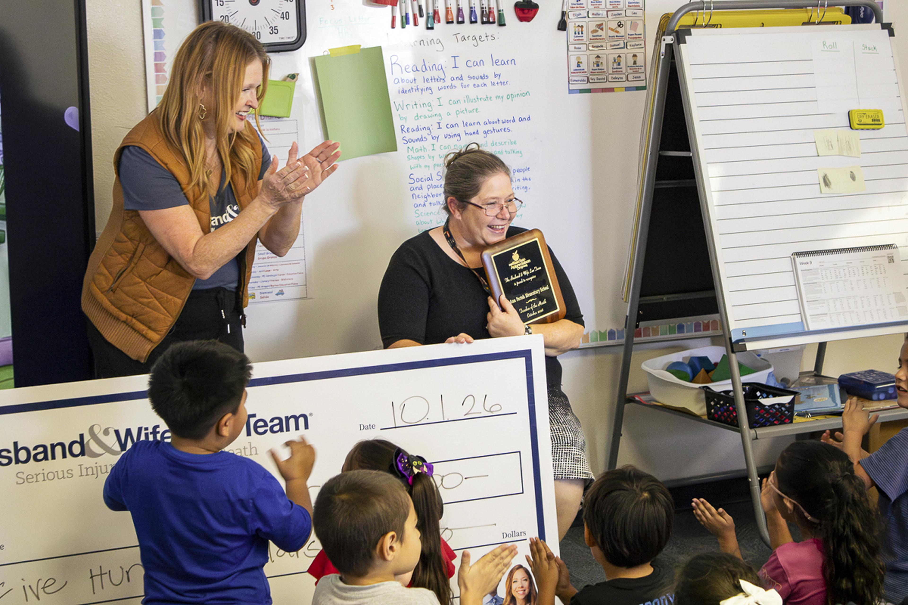 Cheers and applause fill the classroom as Mrs. Warren celebrates her Teacher of the Month honor, surrounded by excited students and Lisa from the Husband & Wife Law Team.