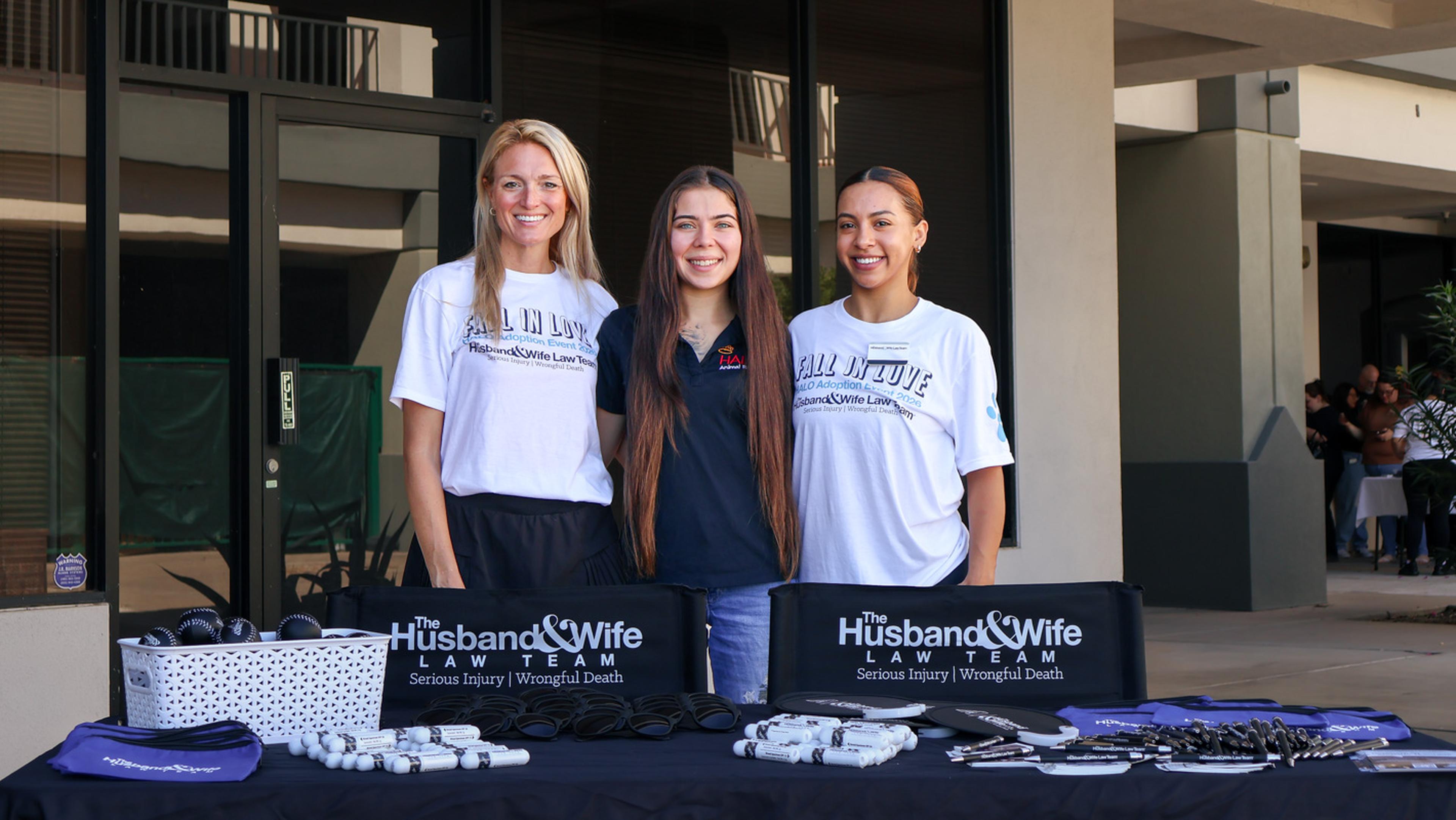 Three Husband & Wife Law Team volunteers stand proudly behind an outreach table, welcoming community members and supporting pet adoption efforts at a HALO Animal Rescue event.