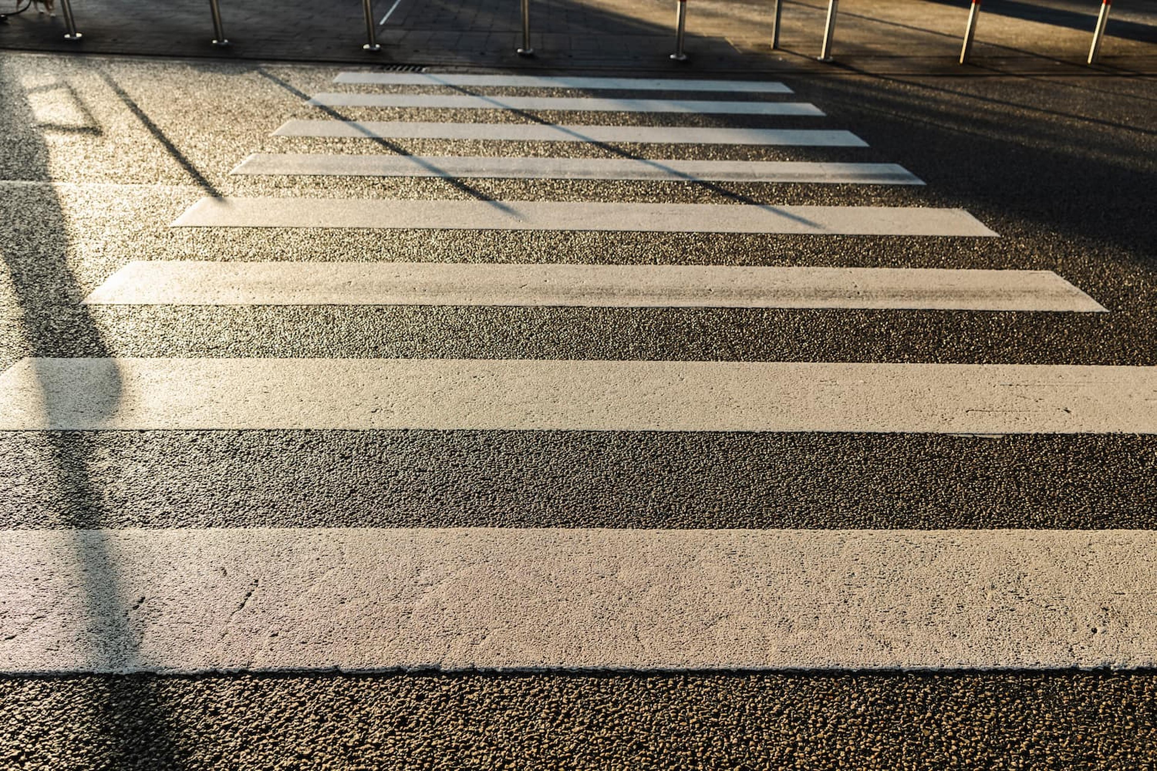 White crosswalk stripes on a city street in late-afternoon sunlight, with long shadows from nearby posts