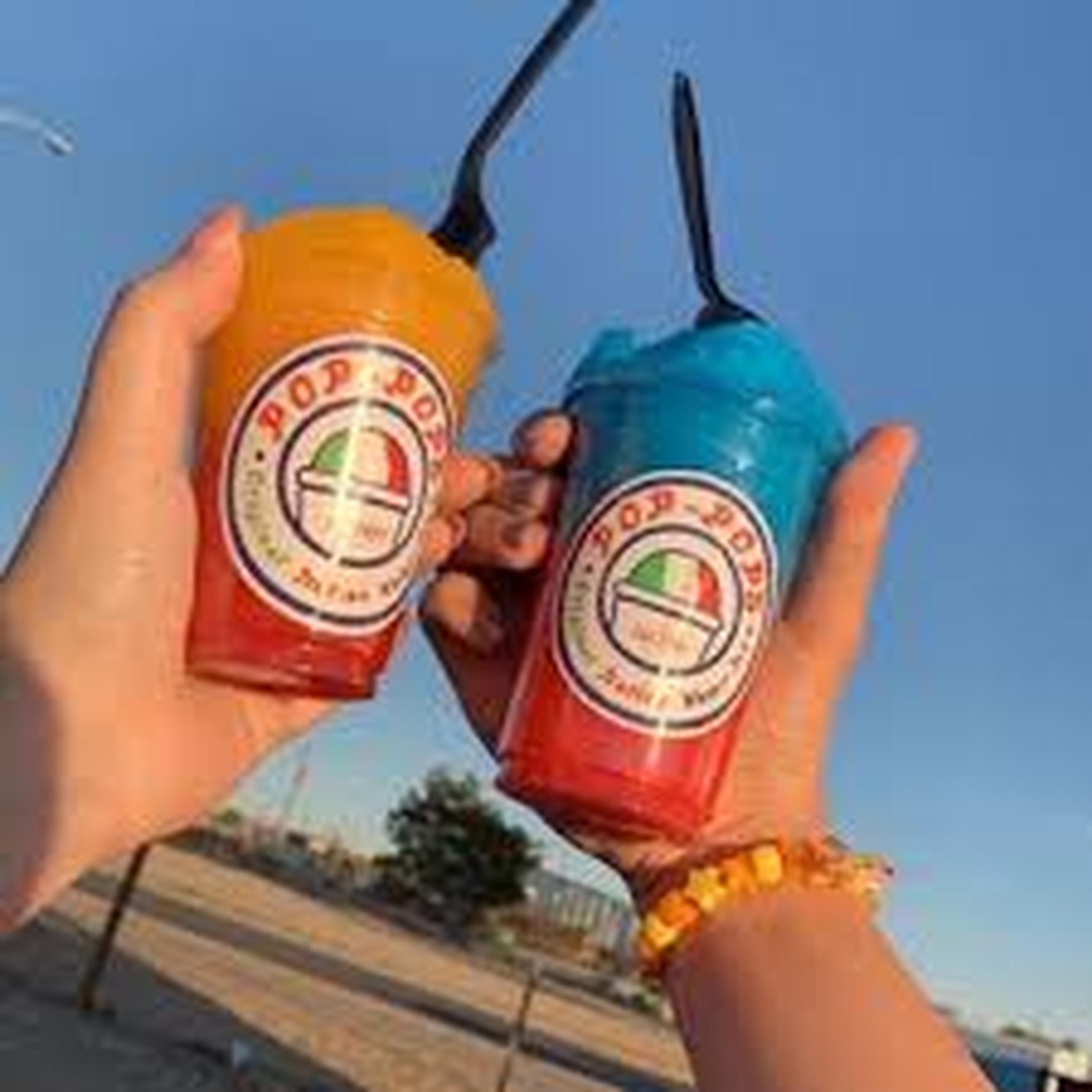 Two hands holding colorful shaved ice drinks in branded cups against a clear blue sky, with a sunny outdoor background.