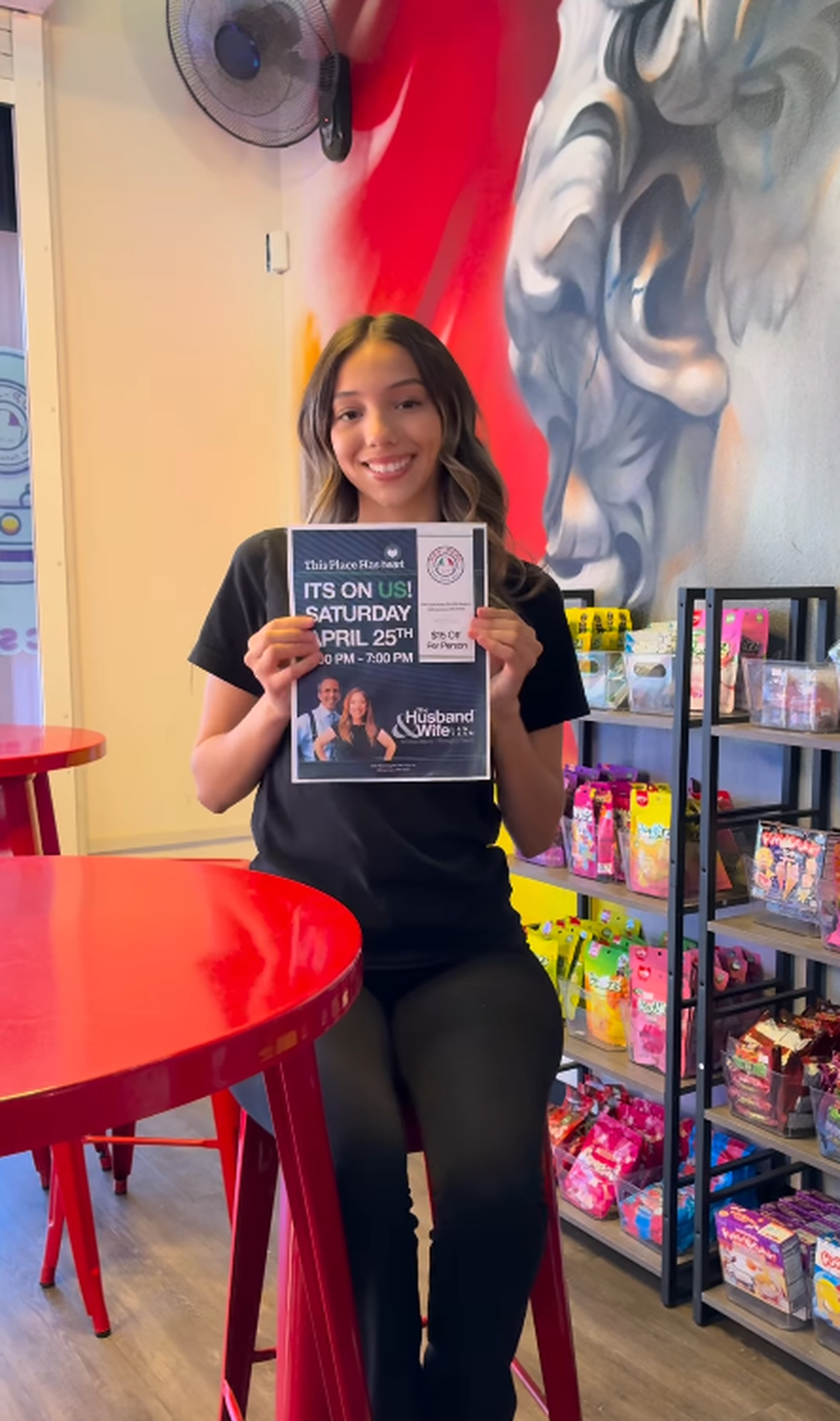 Smiling woman holding a flyer for an event at a candy store, sitting at a red table with shelves of snacks behind her.