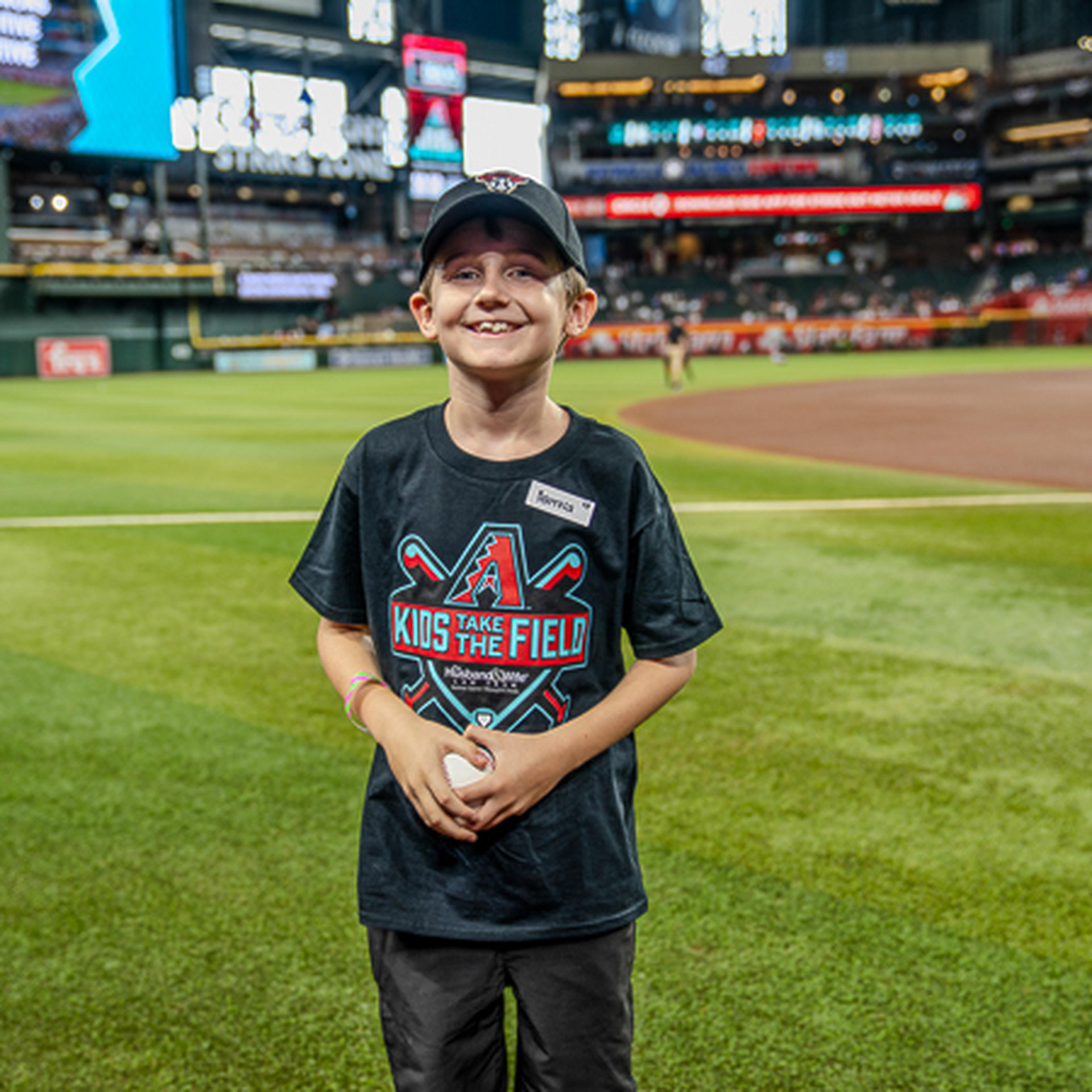 A smiling boy stands on a baseball field wearing a "Kids Take the Field" t-shirt and cap, with a stadium and audience in the background.