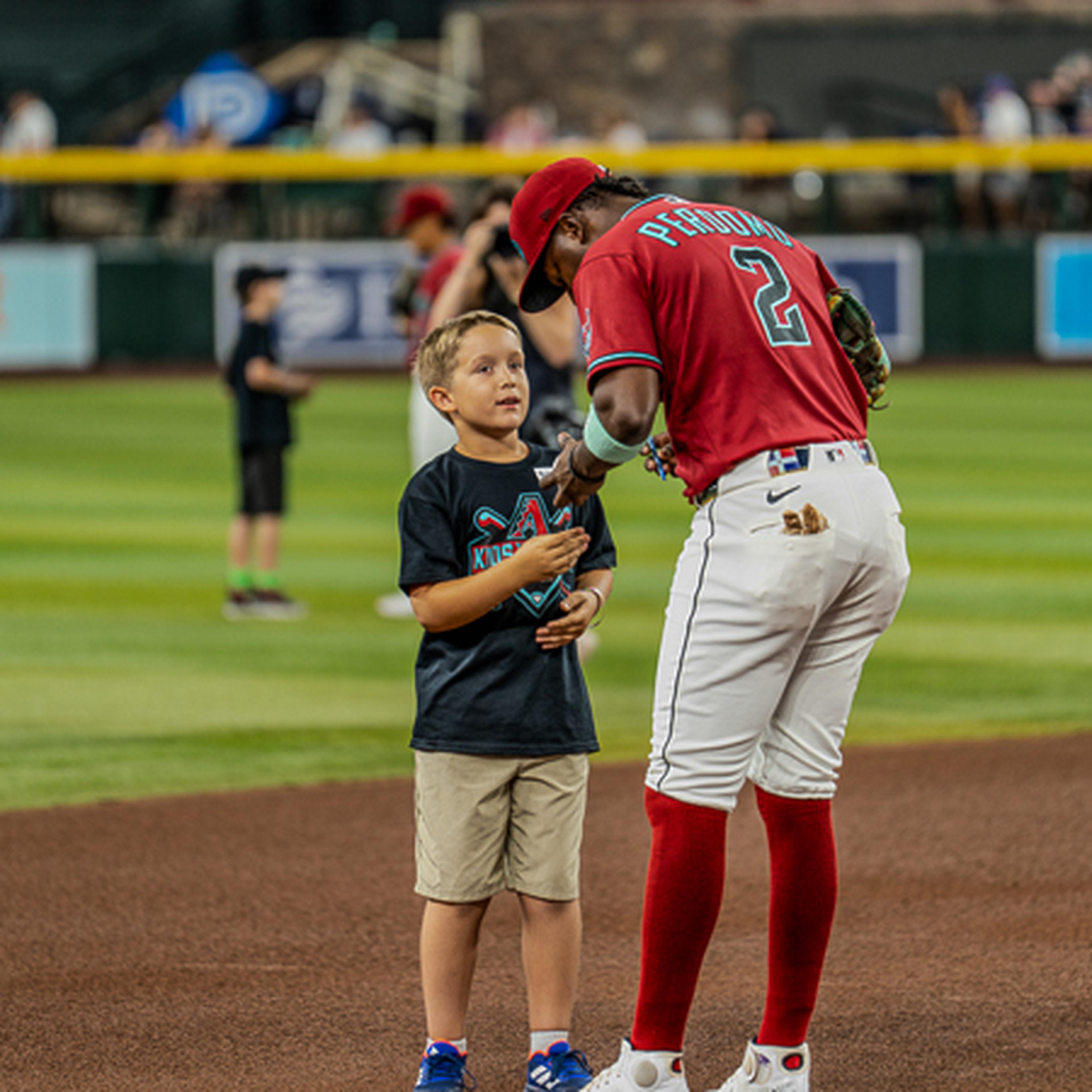 Baseball player in red uniform and cap signs an autograph for a young boy on the field. Other people are in the background.