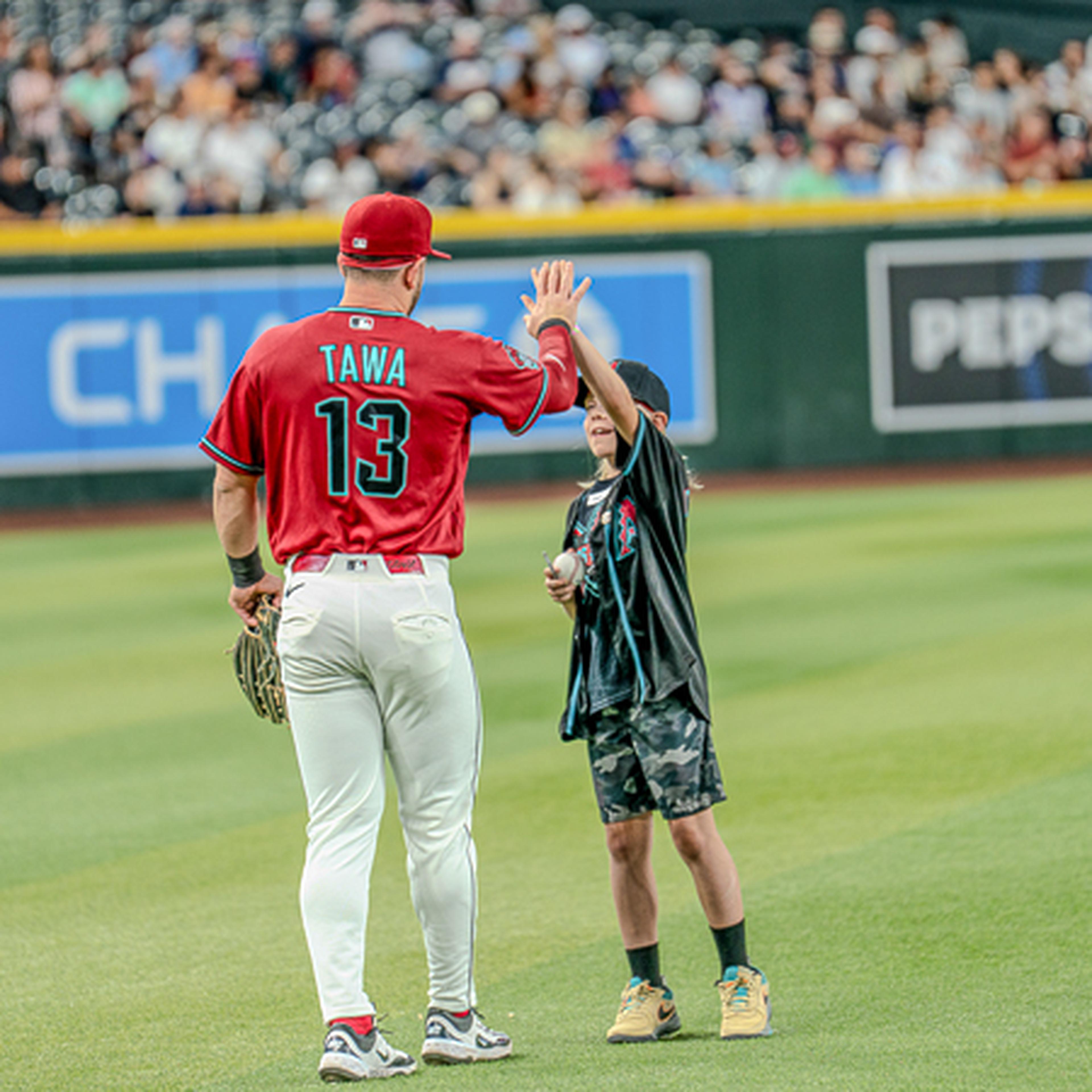 Baseball player in a red jersey high-fives a young fan on the field, with spectators in the background.