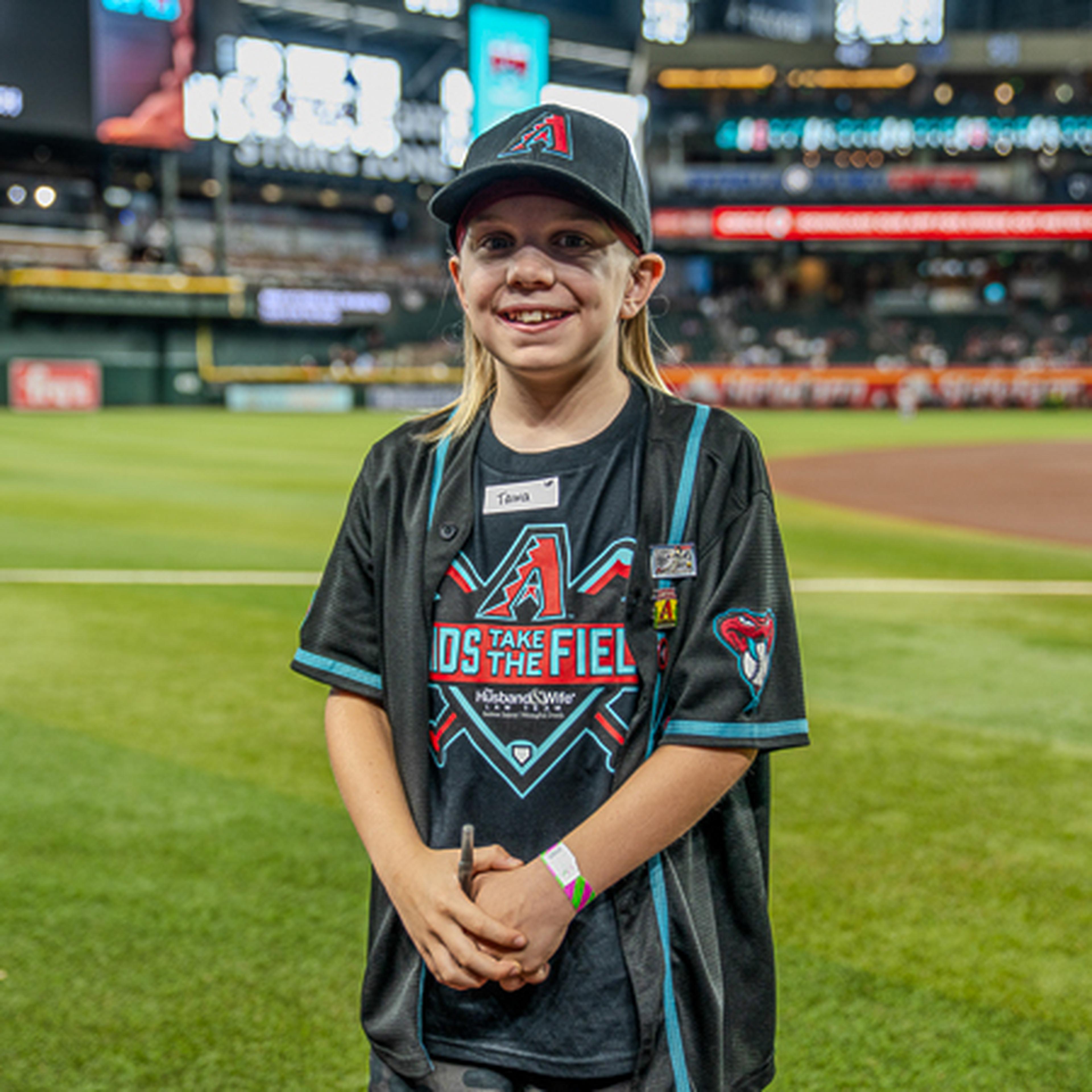 Young fan in a baseball cap and jersey smiles on a stadium field. Stadium seats and lights are visible in the background.