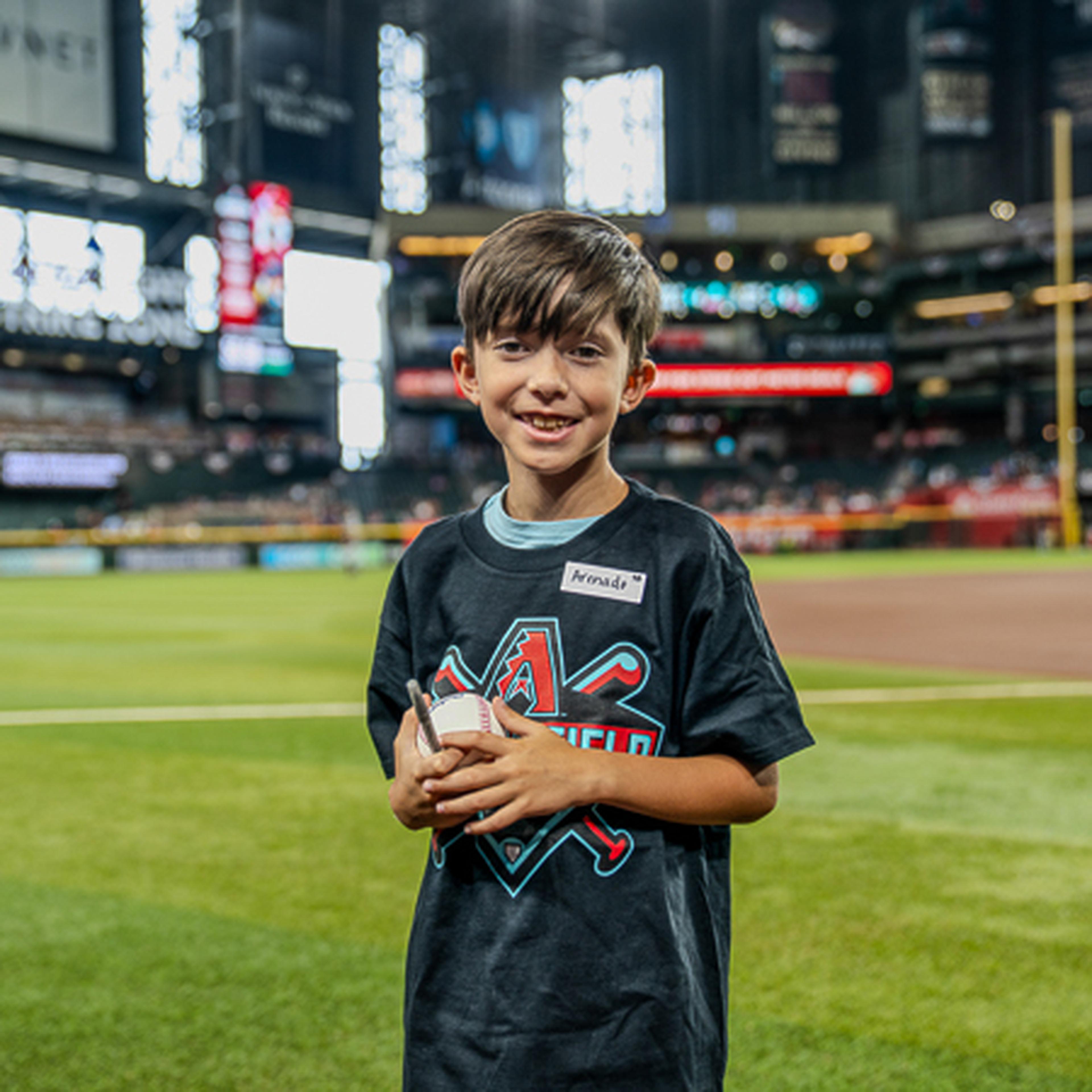Young boy holding a baseball, standing on a grassy field in a stadium, wearing a black Arizona-themed shirt.