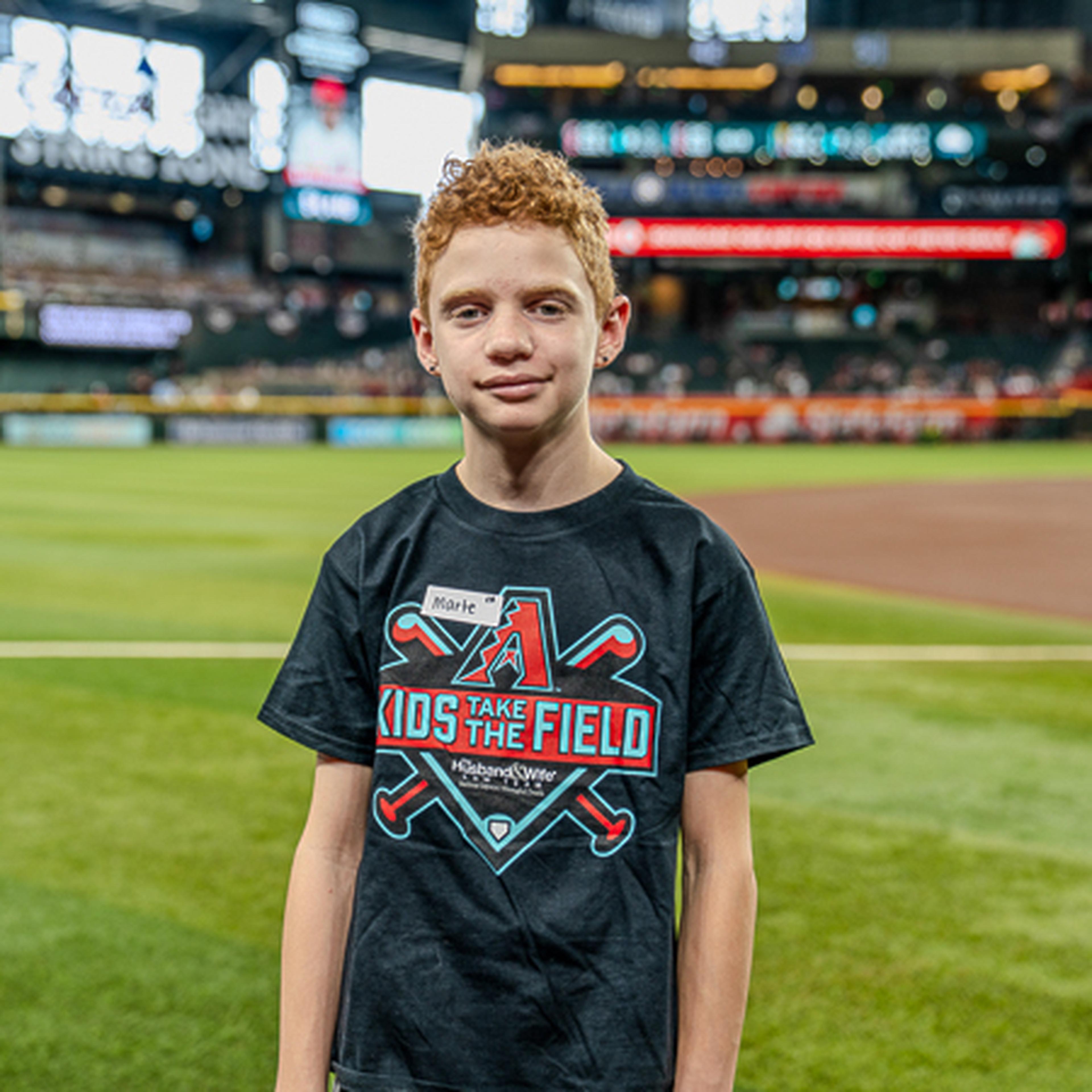 A young person with curly hair standing on a baseball field, wearing a "Kids Take the Field" shirt, in a bustling stadium environment.