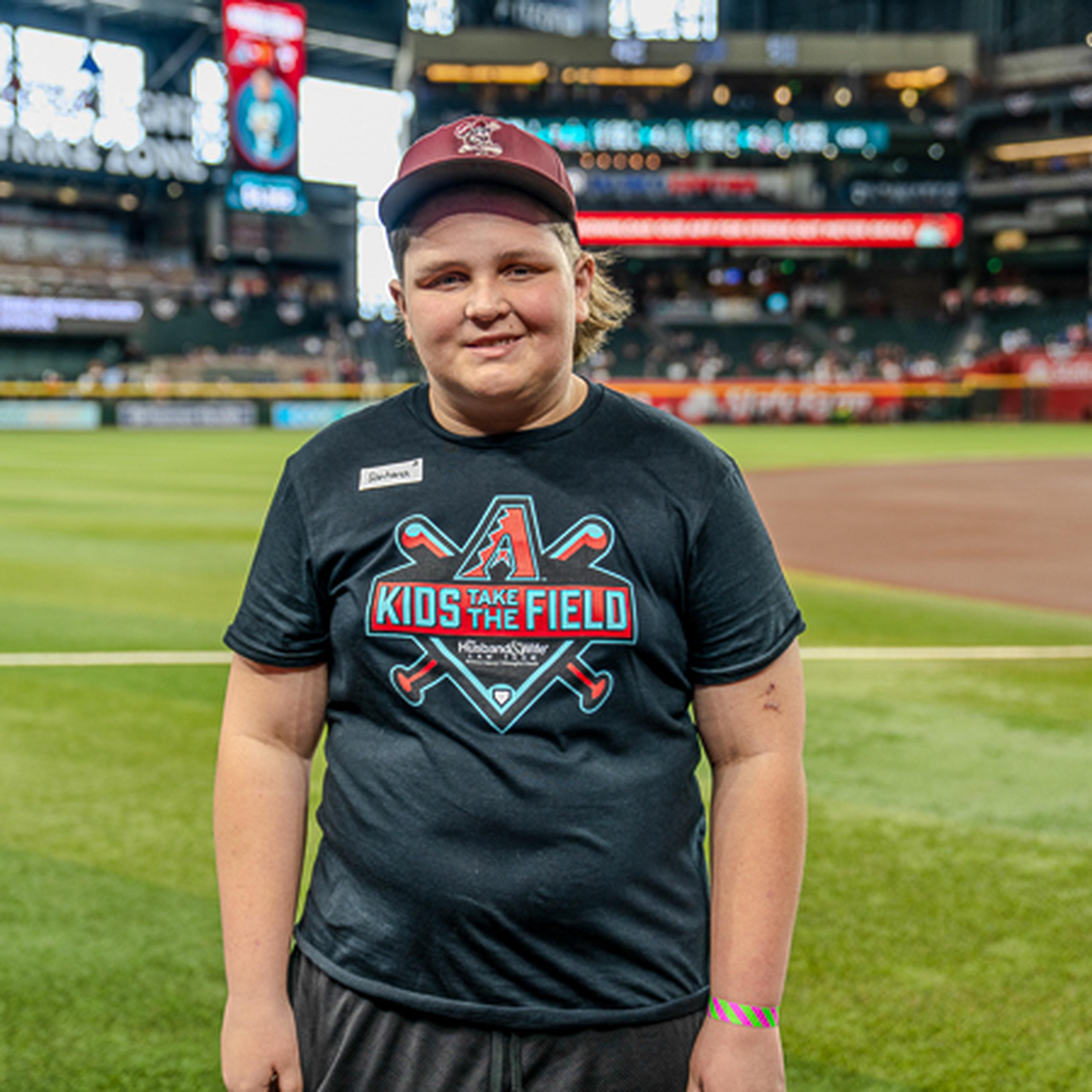 A smiling person in a cap and "Kids Take the Field" t-shirt stands on a baseball field, with a stadium and audience in the background.