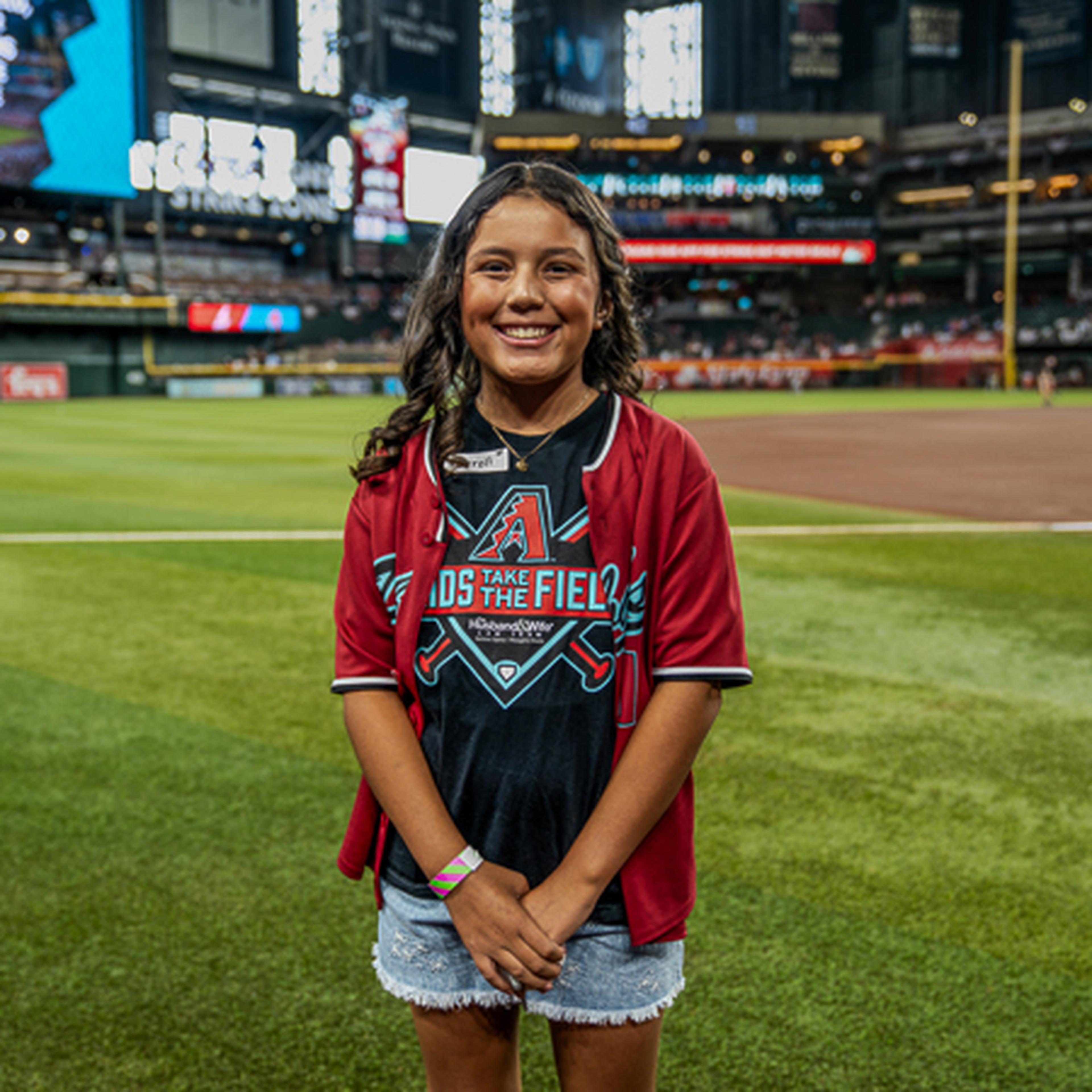Girl smiling on a baseball field, wearing a red jersey over a themed T-shirt, with a stadium and scoreboard in the background.